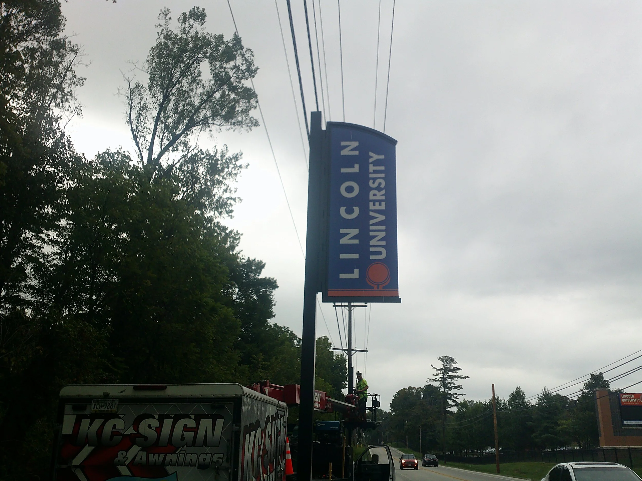 A street scene with a sign for Lincoln University, utility lines overhead, a person in a yellow safety vest working on or near power lines on a bucket lift, and vehicles on the road. The sky is overcast.