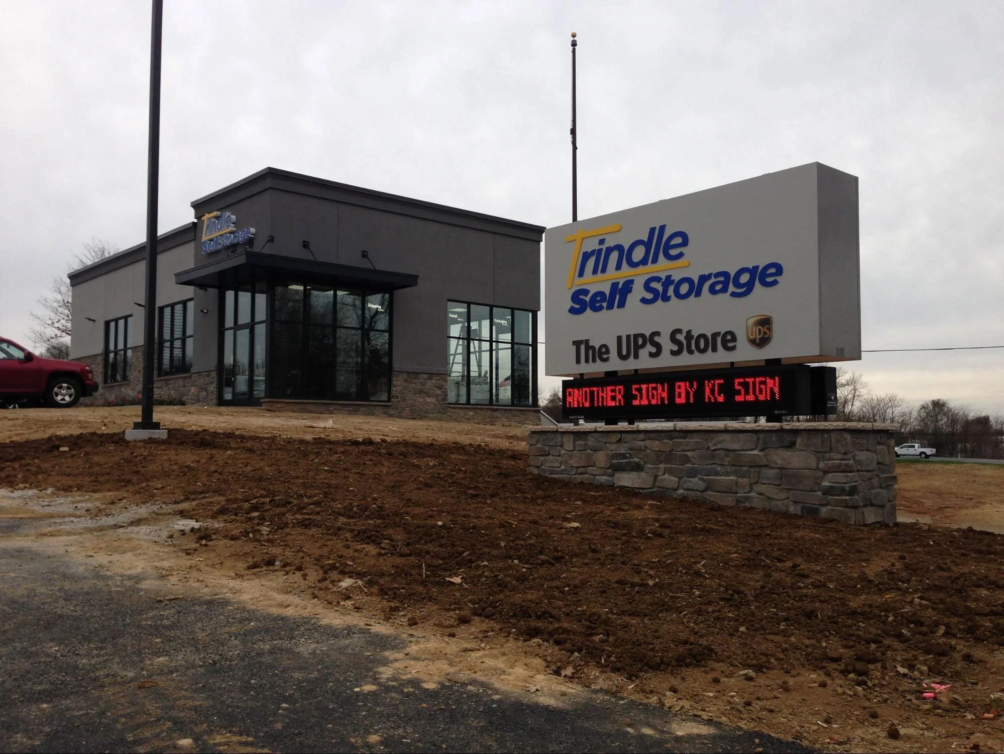 The exterior of a Trindle Self Storage building with a large sign and a digital message board, parked cars, and a cloudy sky in the background.