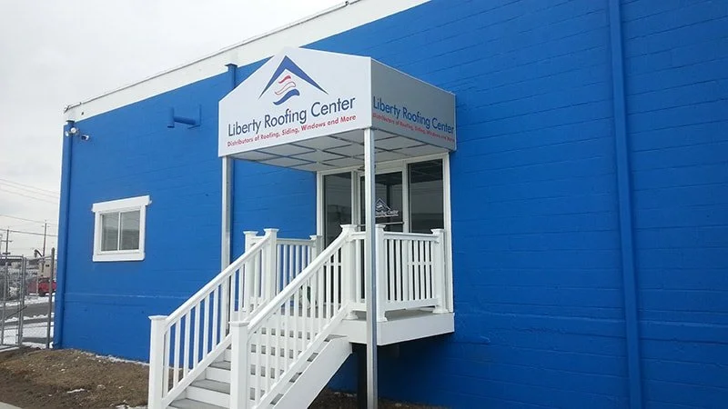 Blue building with a white door and window, featuring a small covered porch with white stairs and railing. Sign above the porch reads Liberty Roofing Center.