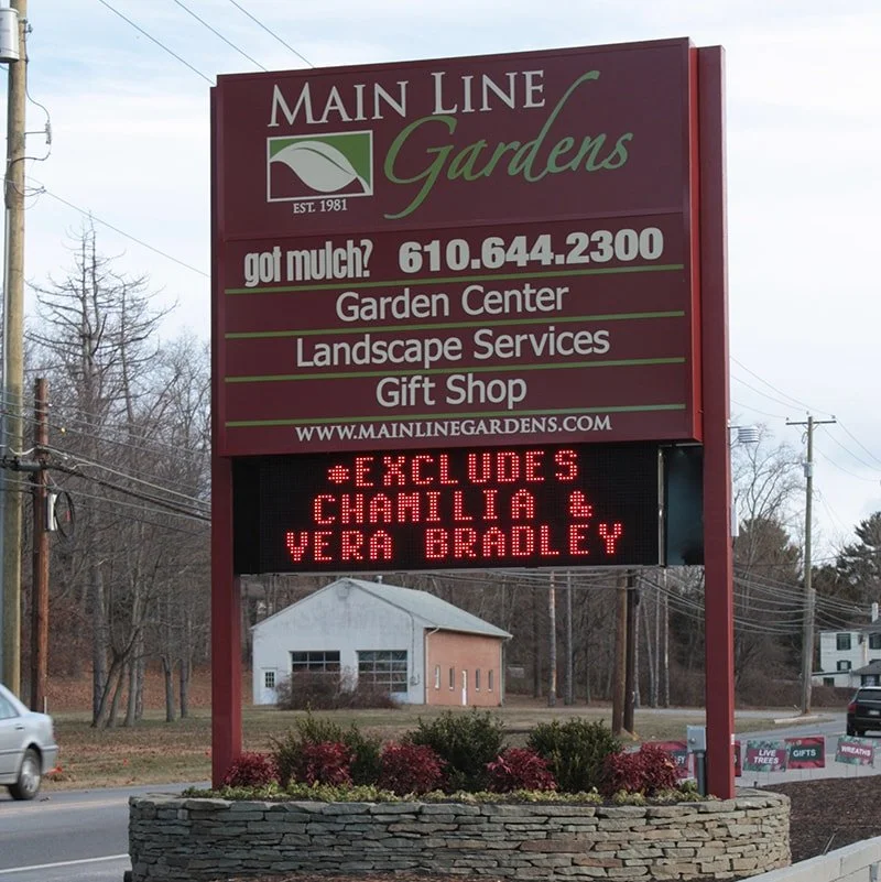 Sign for Main Line Gardens advertising garden and landscape services, with a digital message board below stating 'EXCLUDES CHAMILIA & VERA BRADLEY' in red lights, and a background of trees and buildings.