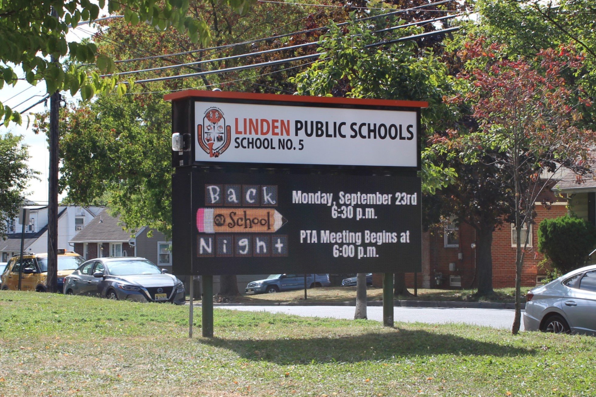 A digital and printed sign outside Linden Public Schools School No. 5, displaying information about the upcoming back-to-school date, PTA meeting time, and other school events, with surrounding trees and parked cars in the background.