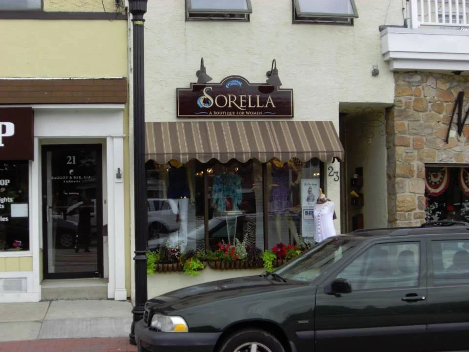 Storefront for Sorella boutique for women with display window, mannequins, and flower arrangements outside, with a black car parked in front.