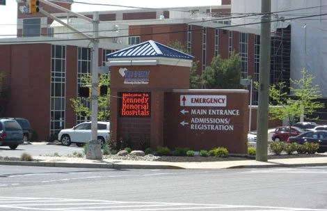 Signboard for Kennedy Memorial Hospital with directions to emergency, main entrance, and admissions/registration, in an urban setting.