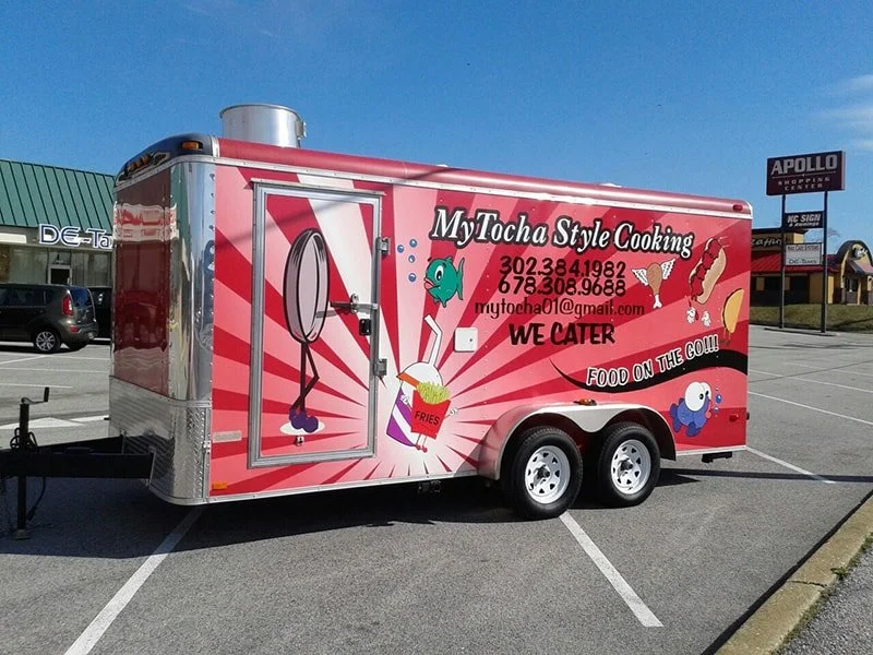 Pink food trailer with cartoon fish, fried foods, and food items, advertising My Tocha Style Cooking with contact numbers and email, in a parking lot with a shopping center in the background.