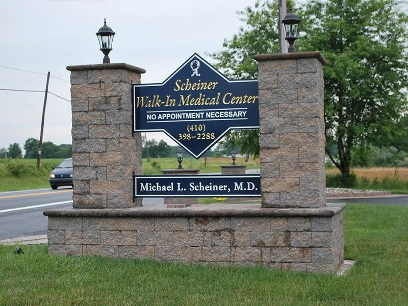 Sign for Scheiner Walk-In Medical Center, operated by Dr. Michael L. Scheiner, M.D., with contact information, situated between two stone pillars with street and greenery in the background.