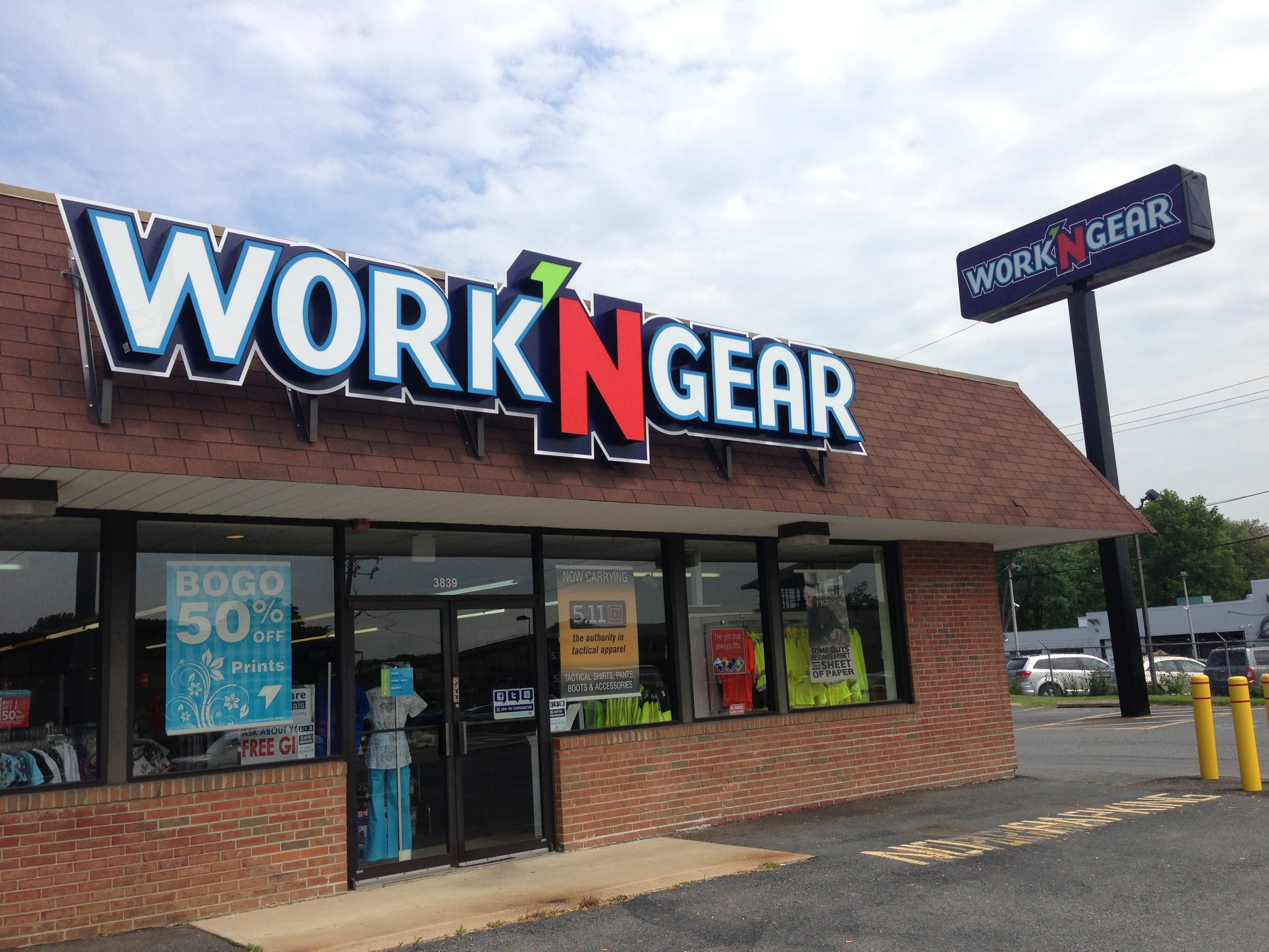 A storefront with a large sign reading 'WORK 'N GEAR' on the building and a smaller sign on a pole with the same name. The store sells tactical shirts, pants, boots, and accessories, and has promotional posters in the window.