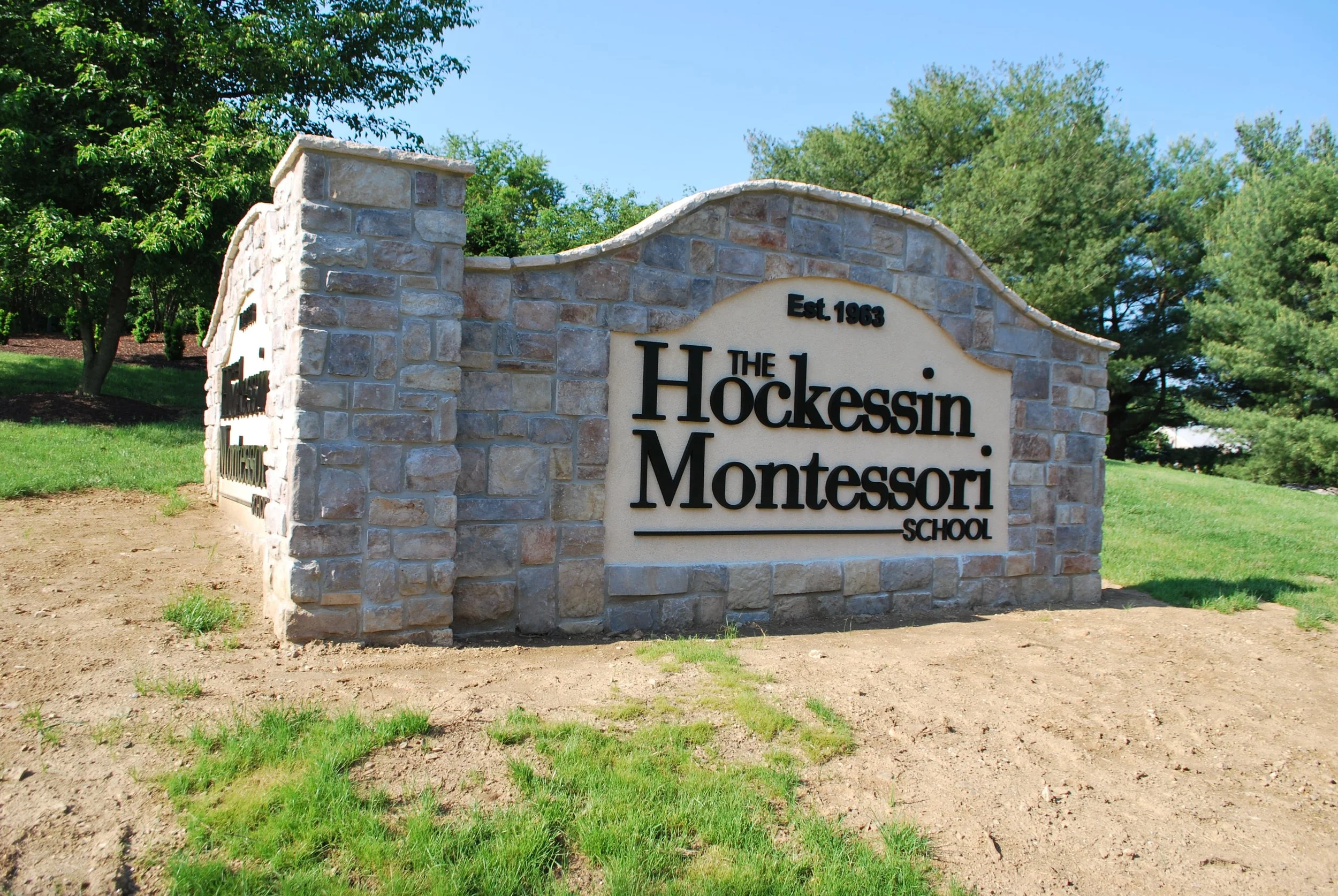 Entrance sign for Hockessin Montessori School, established in 1963, made of stone with black lettering, placed on a patch of dirt with green grass and trees in the background.