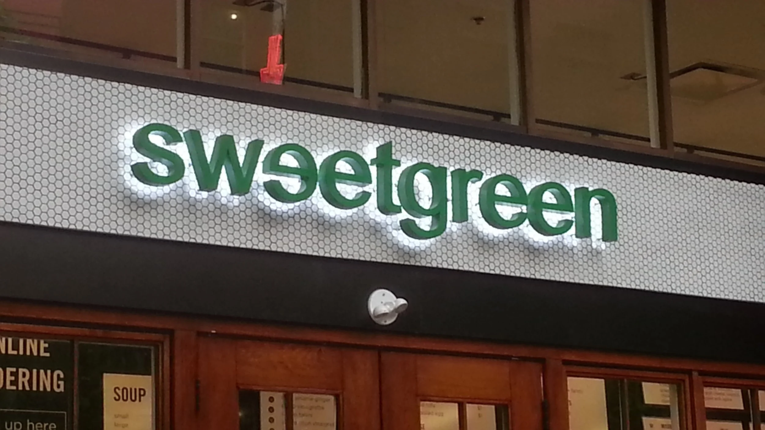 Indoor sign displaying the store name 'Sweetgreen' in green 3D letters, mounted on a white background with a honeycomb pattern.