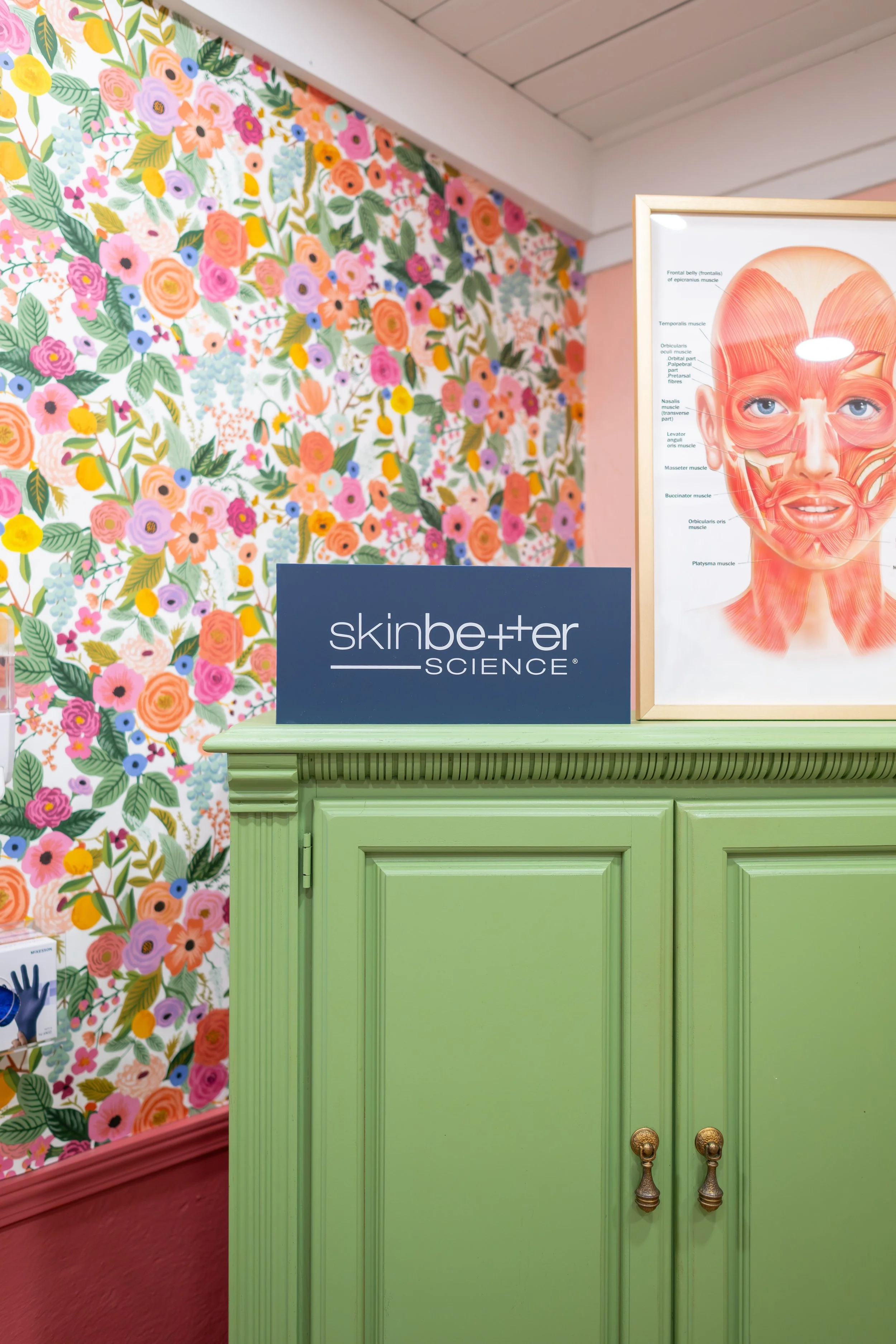 Green cabinet with a SkinBetter Science sign and a diagram of facial muscles on top, with Rifle Paper pink floral wallpaper in the background.
