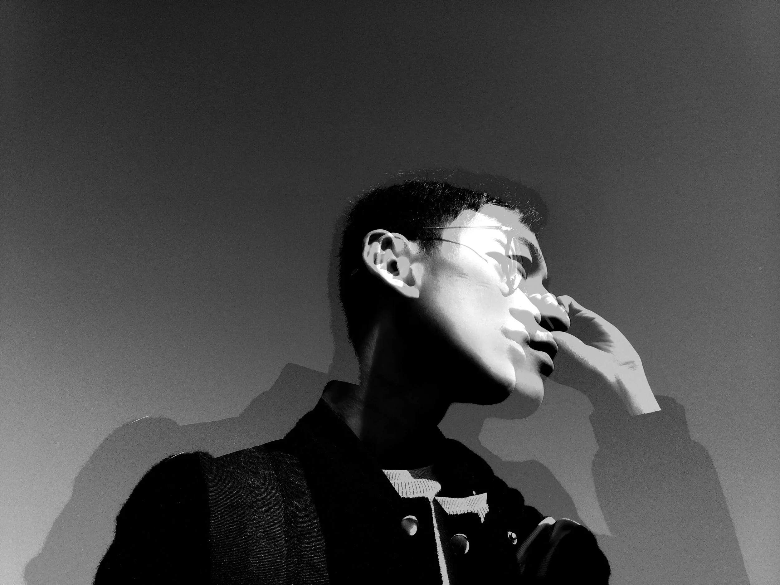Black and white photo of a person with short hair and glasses, looking to the side, with their hand near their face across a plain background.