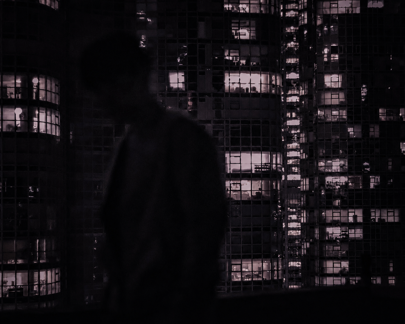 A silhouetted person standing in front of a cityscape at night, with illuminated office building windows in the background.