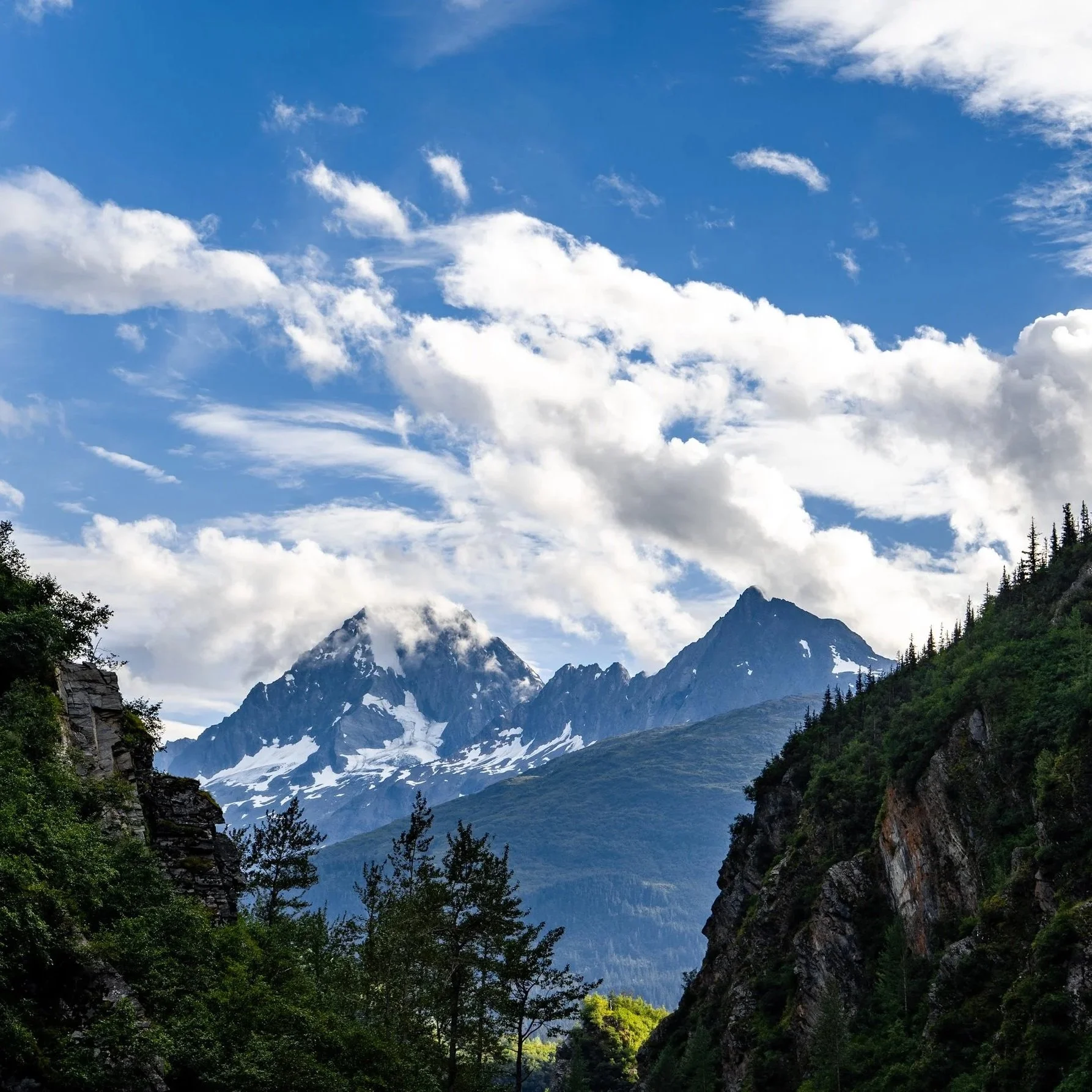 Scenic mountain landscape with snow-capped peaks, green forest, and a partly cloudy sky.