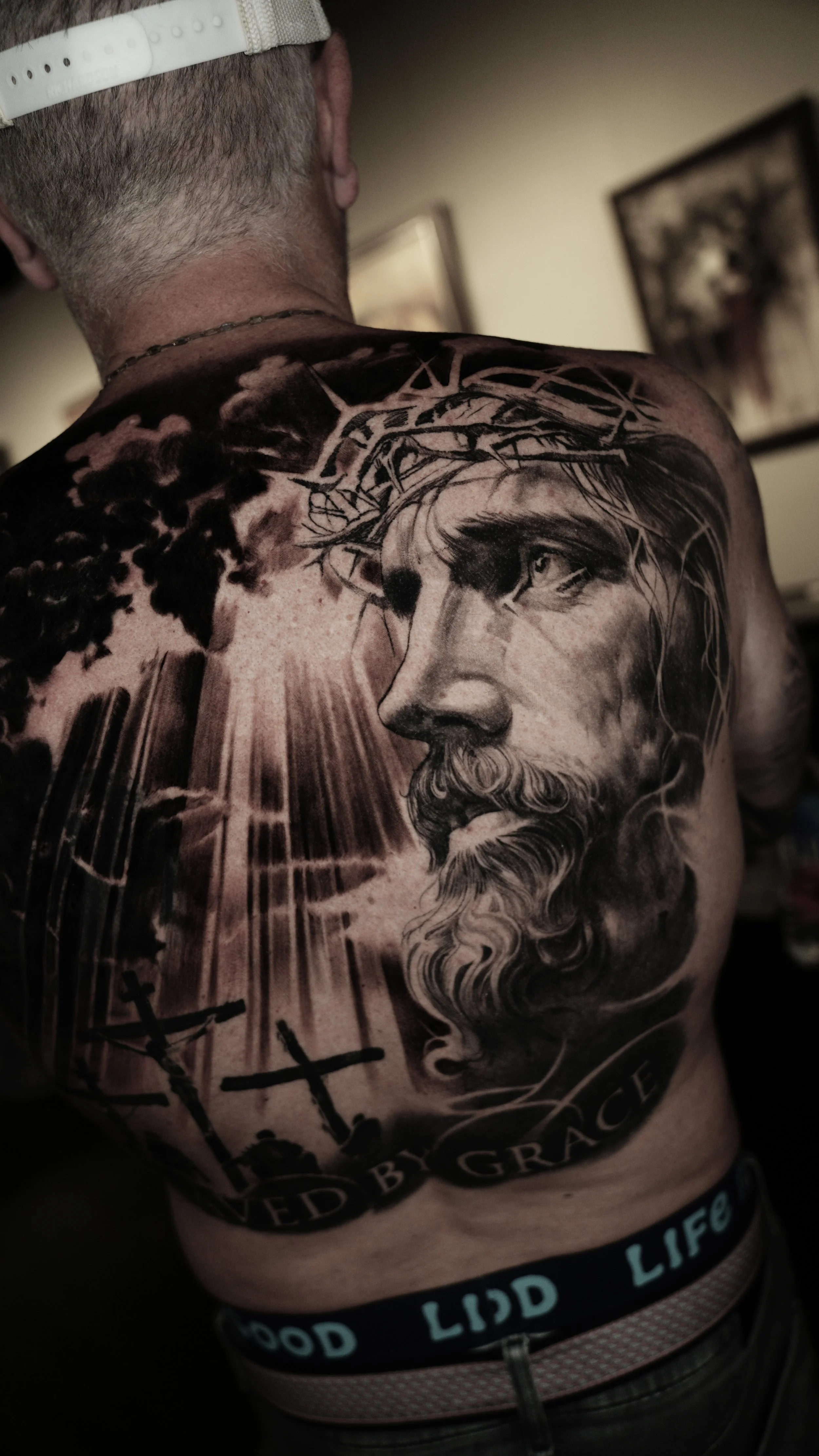 A person's back and shoulder featuring a detailed black and gray tattoo of Jesus Christ with a crown of thorns. The tattoo includes a cross, dark clouds, and rays of light. The person is wearing a black waistband with the text 'Good Lidd Life' in tur