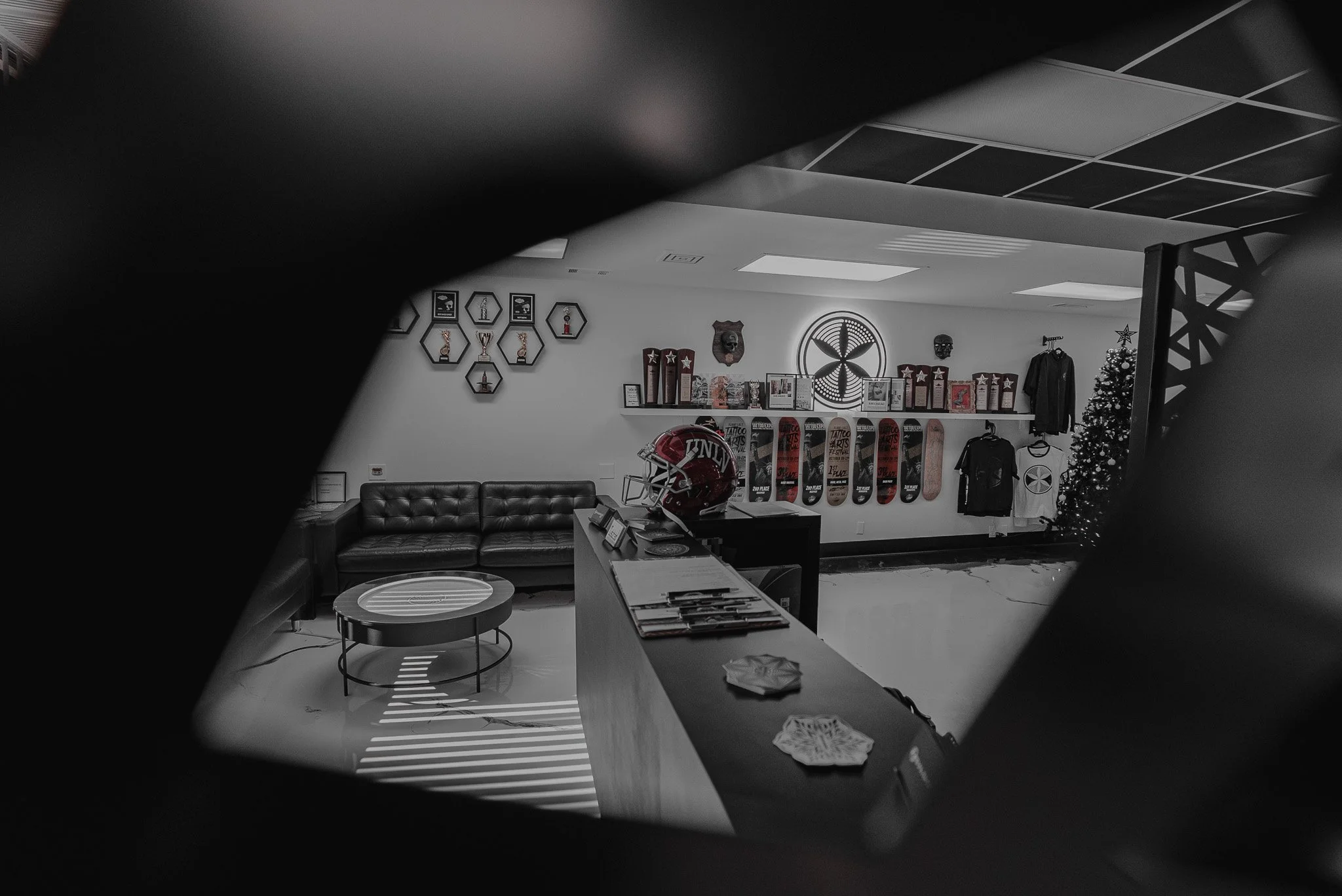 Interior of a sports-themed room with skateboard decks on the wall, trophies, medals, and a football helmet on a desk, with a black leather couch, a round coffee table, a decorated Christmas tree, and wall decorations, viewed through a black framing 