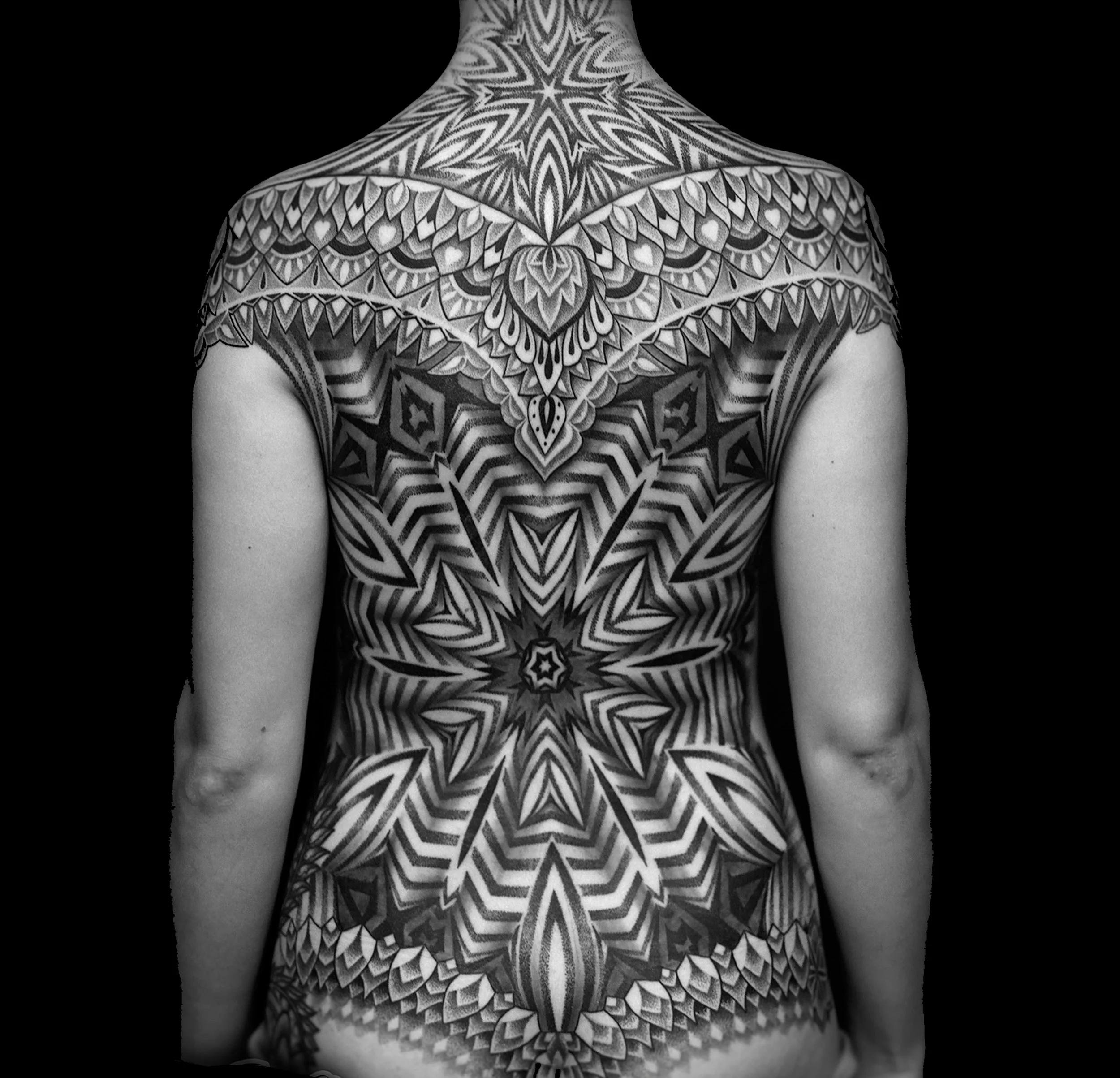 Back of a person with intricate black and white geometric tattoo covering the shoulder, back, and upper arms, set against a black background.