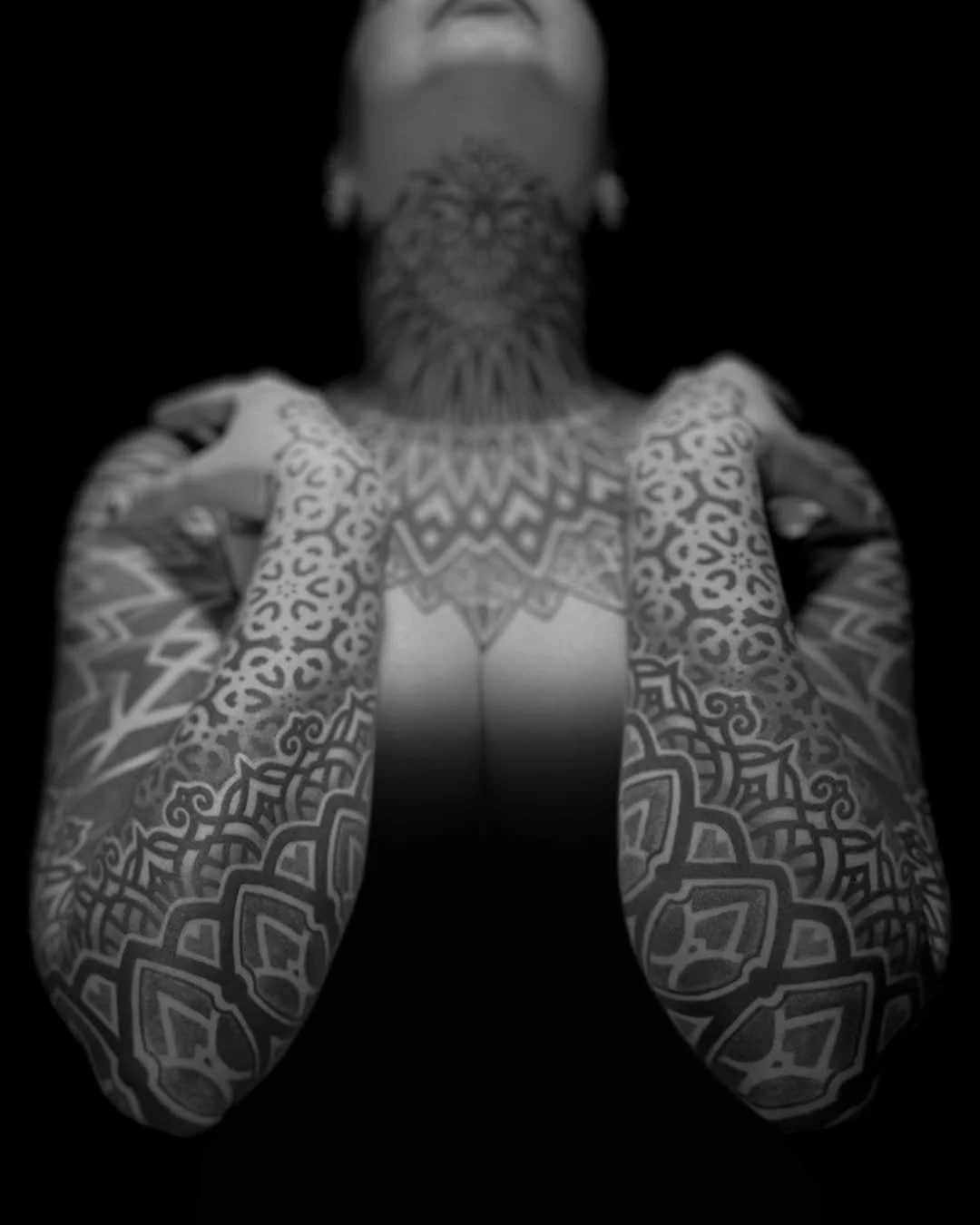 A person with extensive tattoos on their arms, chest, and neck, holding their hands on their shoulders, with face partially visible, in black and white.