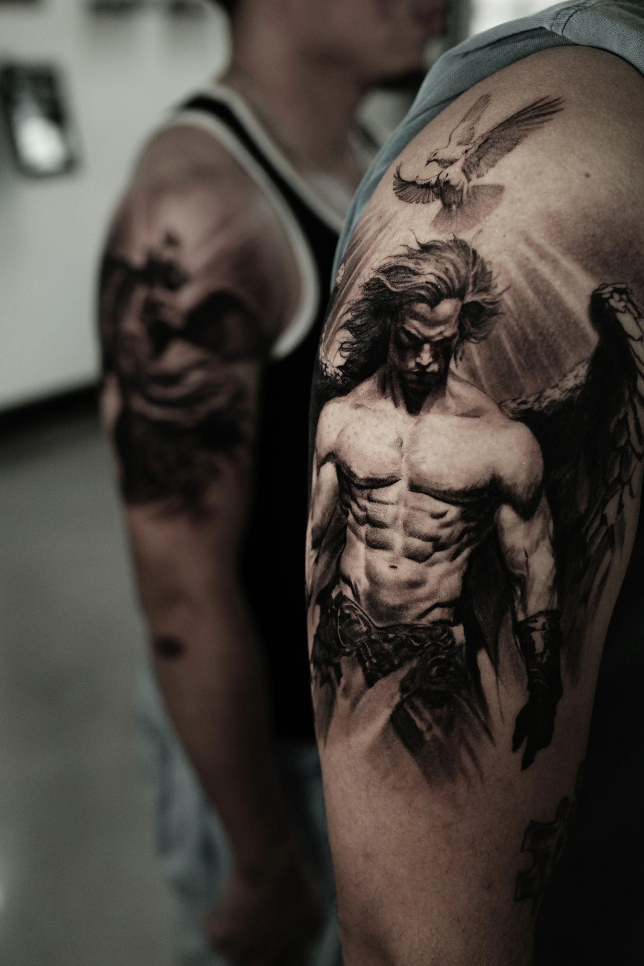 Close-up of a person’s shoulder and upper arm showcasing a detailed black and gray tattoo of a woman with long hair, muscular physique, and a stern expression, with a bird flying above her.