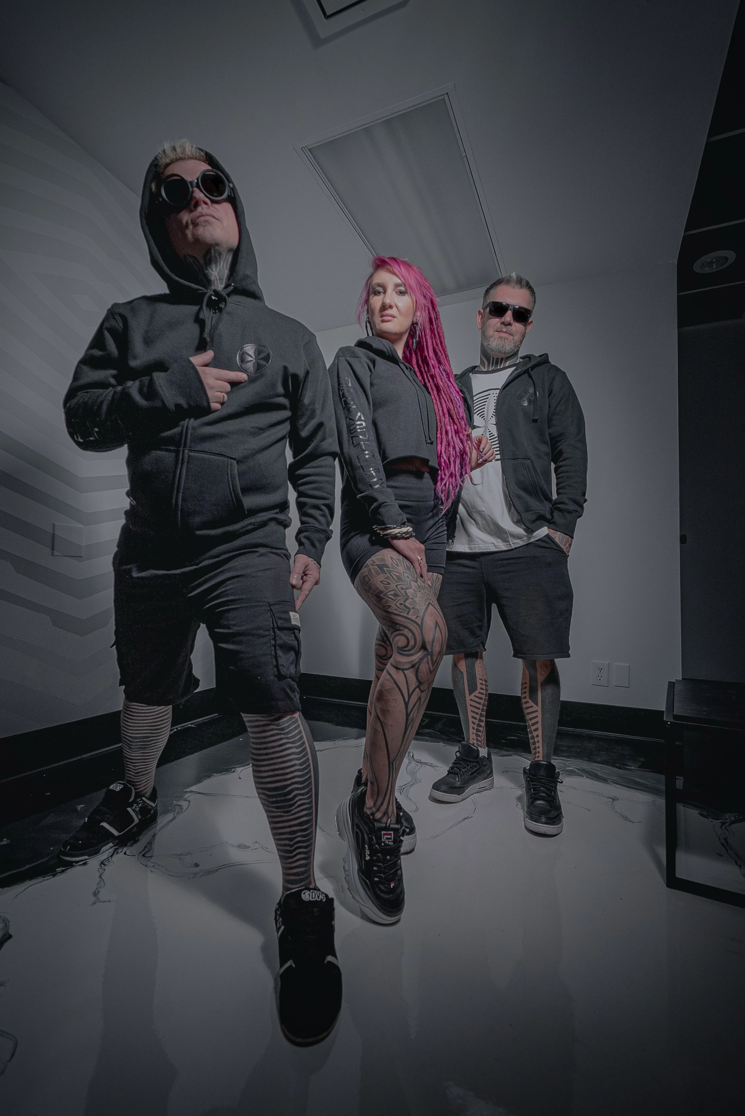 Three people standing indoors with a reflective floor, wearing black hoodies, shorts, and sunglasses, with one woman having pink hair and tattoos on her legs.