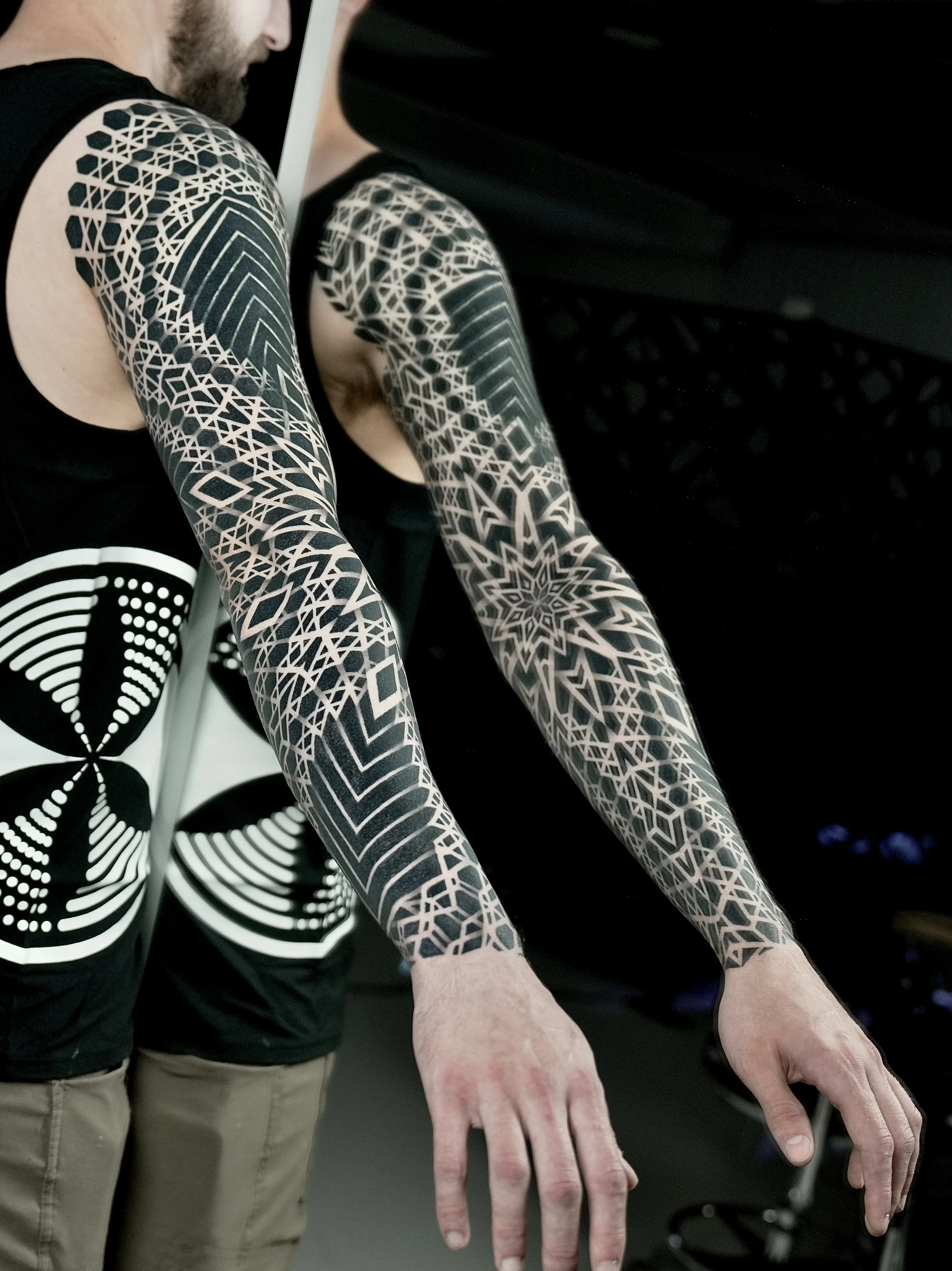 A person with intricate black and white geometric tattoos covering both arms. The person is bending forward with arms extended downward. The individual is wearing a black sleeveless shirt and beige pants, and part of the face and beard are visible.