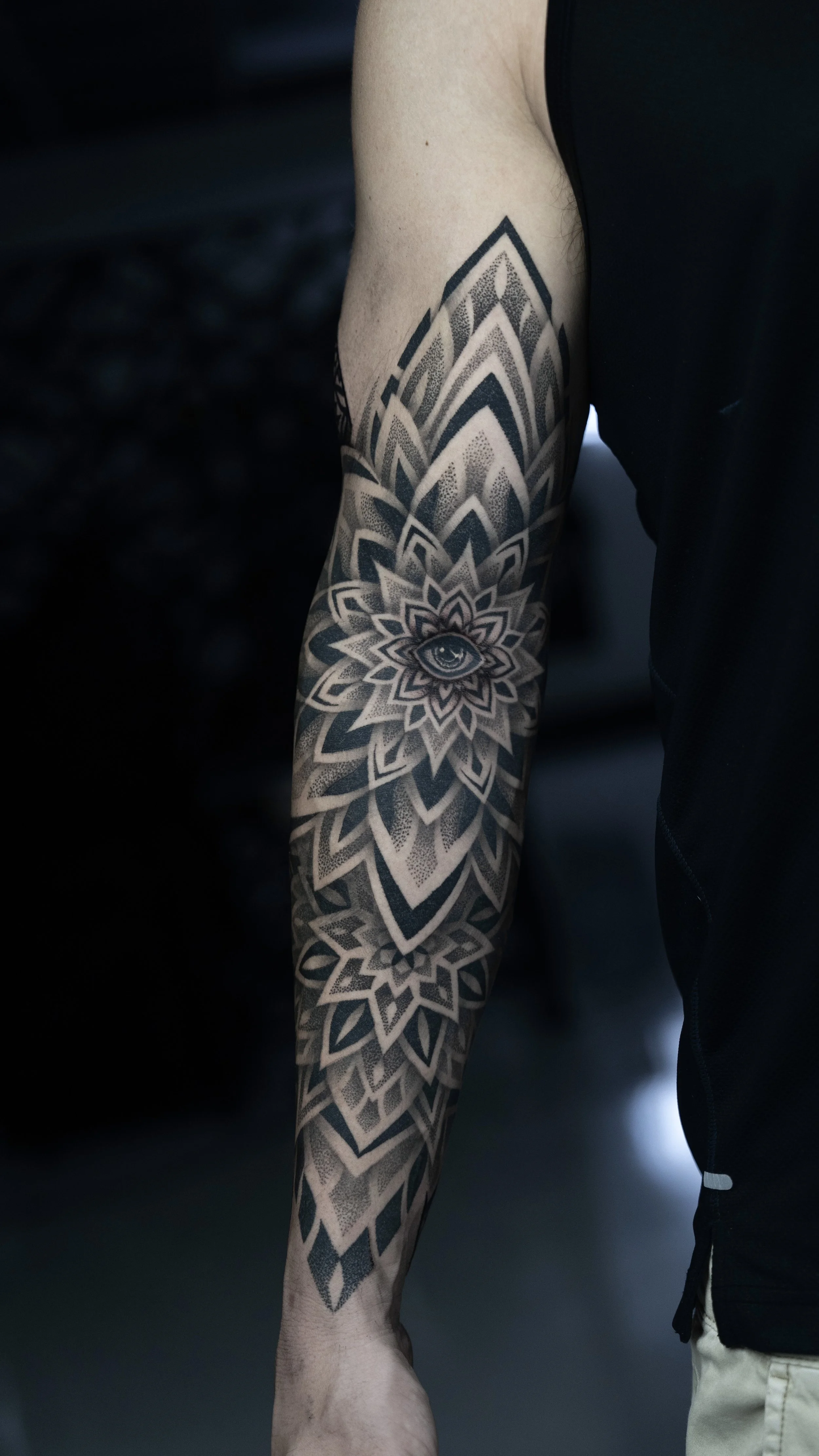 Close-up of a person's arm with a detailed black and grey tattoo featuring an eye at the center of a large geometric flower pattern.