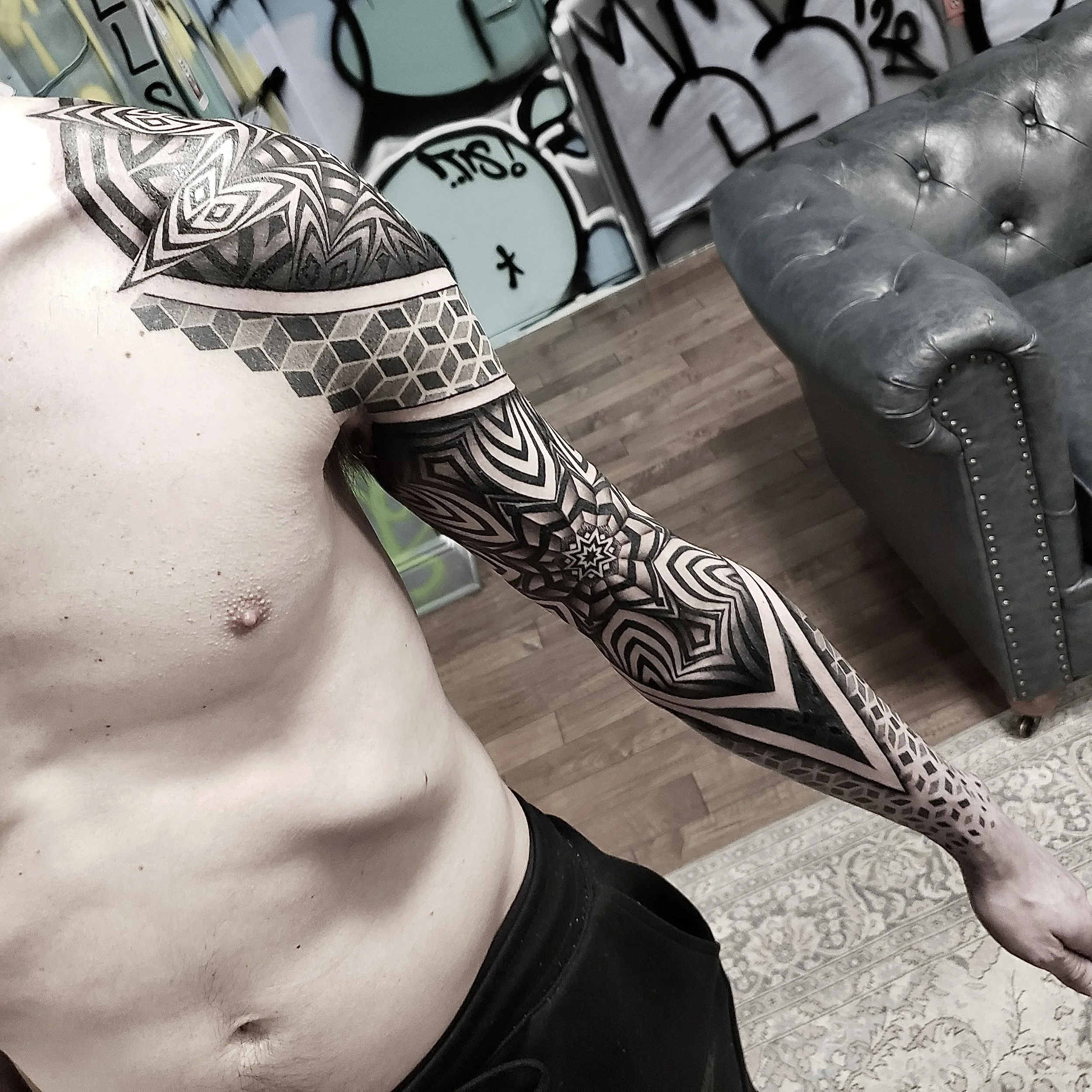 A person with detailed black and gray tattoos on their left arm, shoulder, and chest, standing in a room with graffiti-style art on the walls, a gray leather armchair, and a patterned rug.