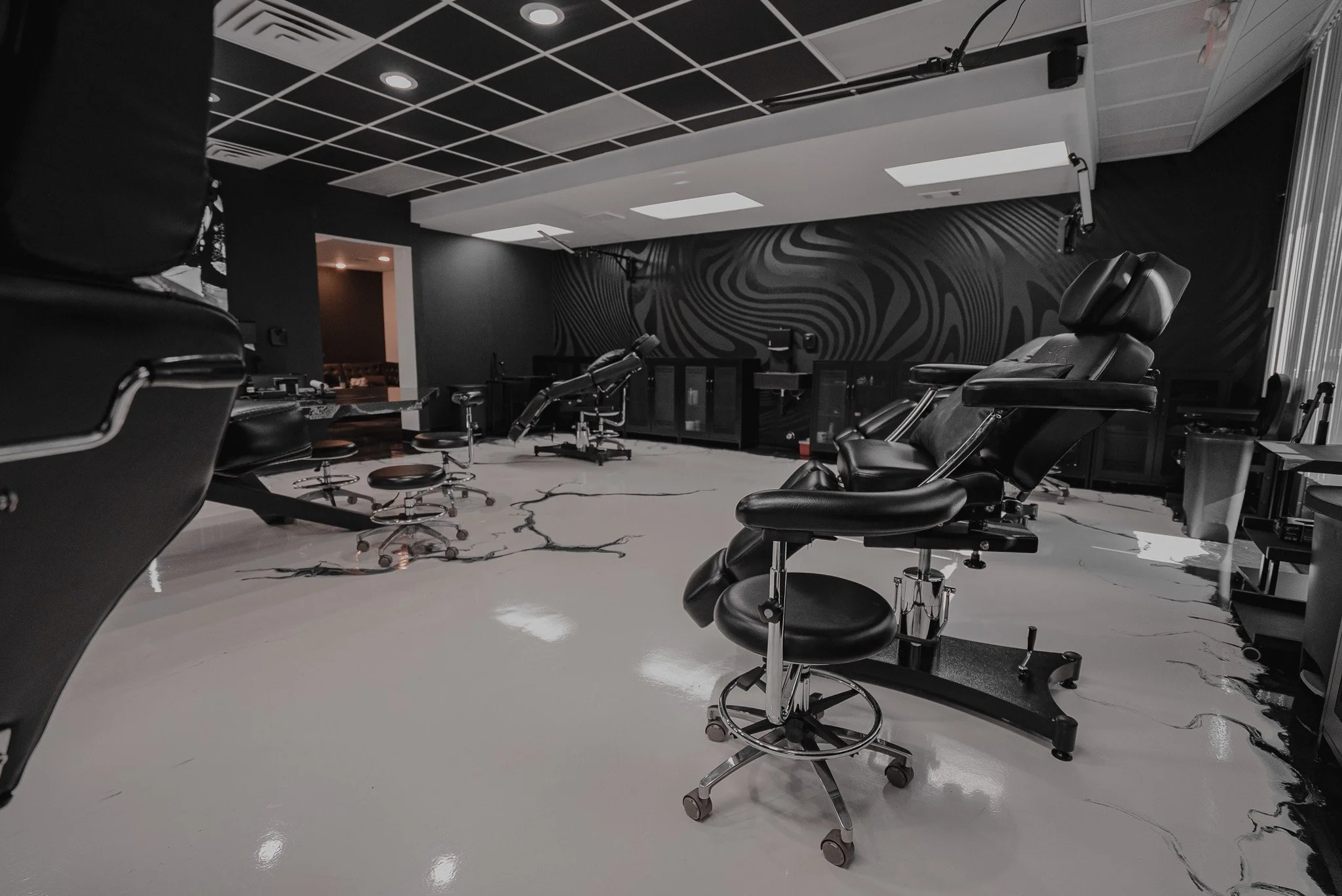A black and white photo of a tattoo studio with tattoo chairs, stools, and equipment, set against a stylish black wall with abstract patterns.