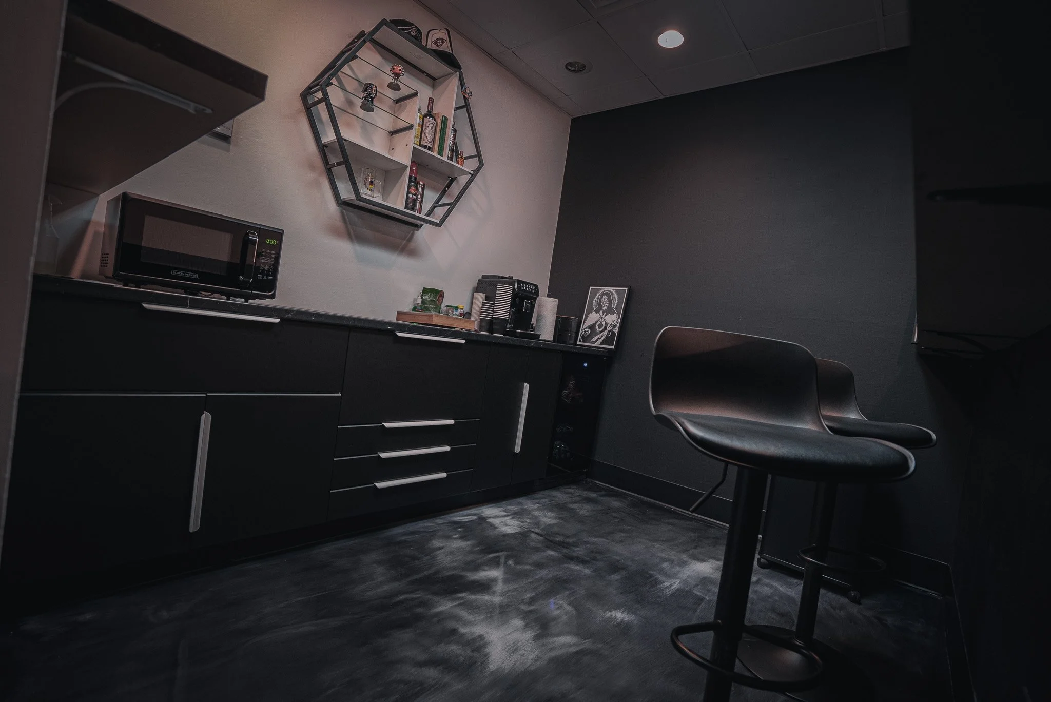 A small kitchenette in a dark room with black cabinets, a microwave, and a countertop with various items including bottles, a framed picture of Jesus, and a coffee machine. Two black bar stools are in front of the counter, and a geometric shelf with 