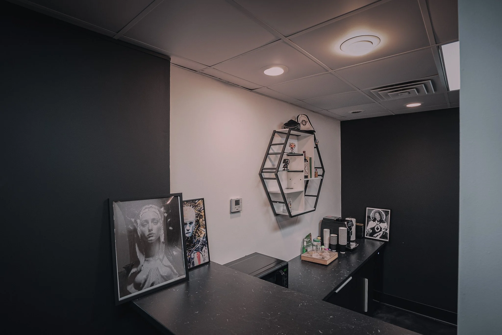 Corner of a room with black and white walls, ceiling lights, a black countertop with framed pictures, a shelf with small decorative items, and a coffee machine area.