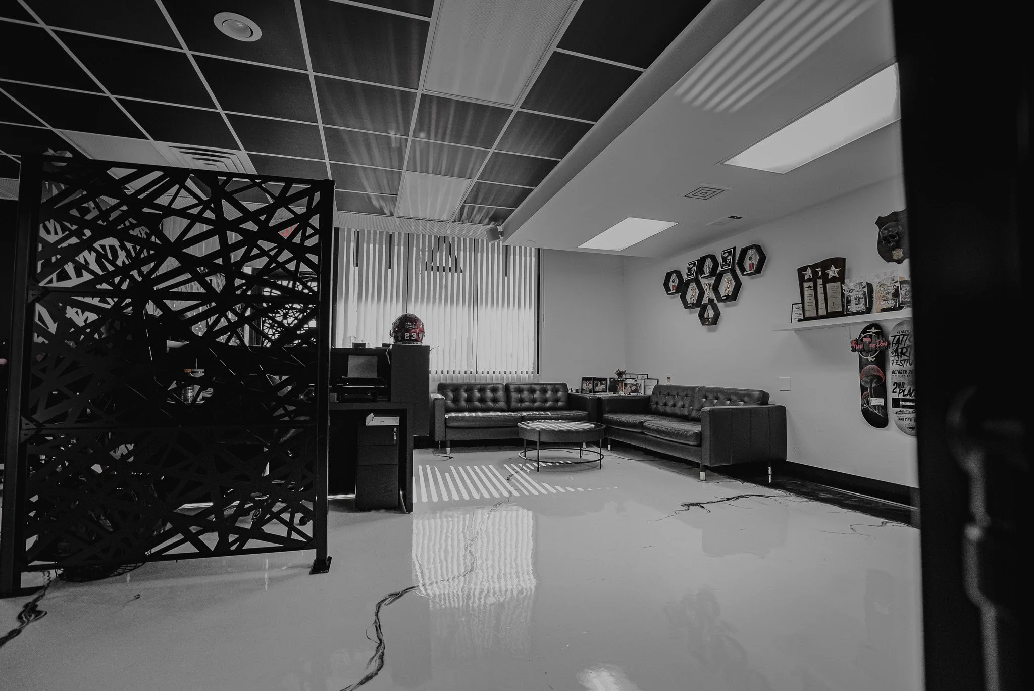 Empty modern lounge with black leather sofas, coffee table, skateboards on the wall, helmet on a stand, and a window with blinds.