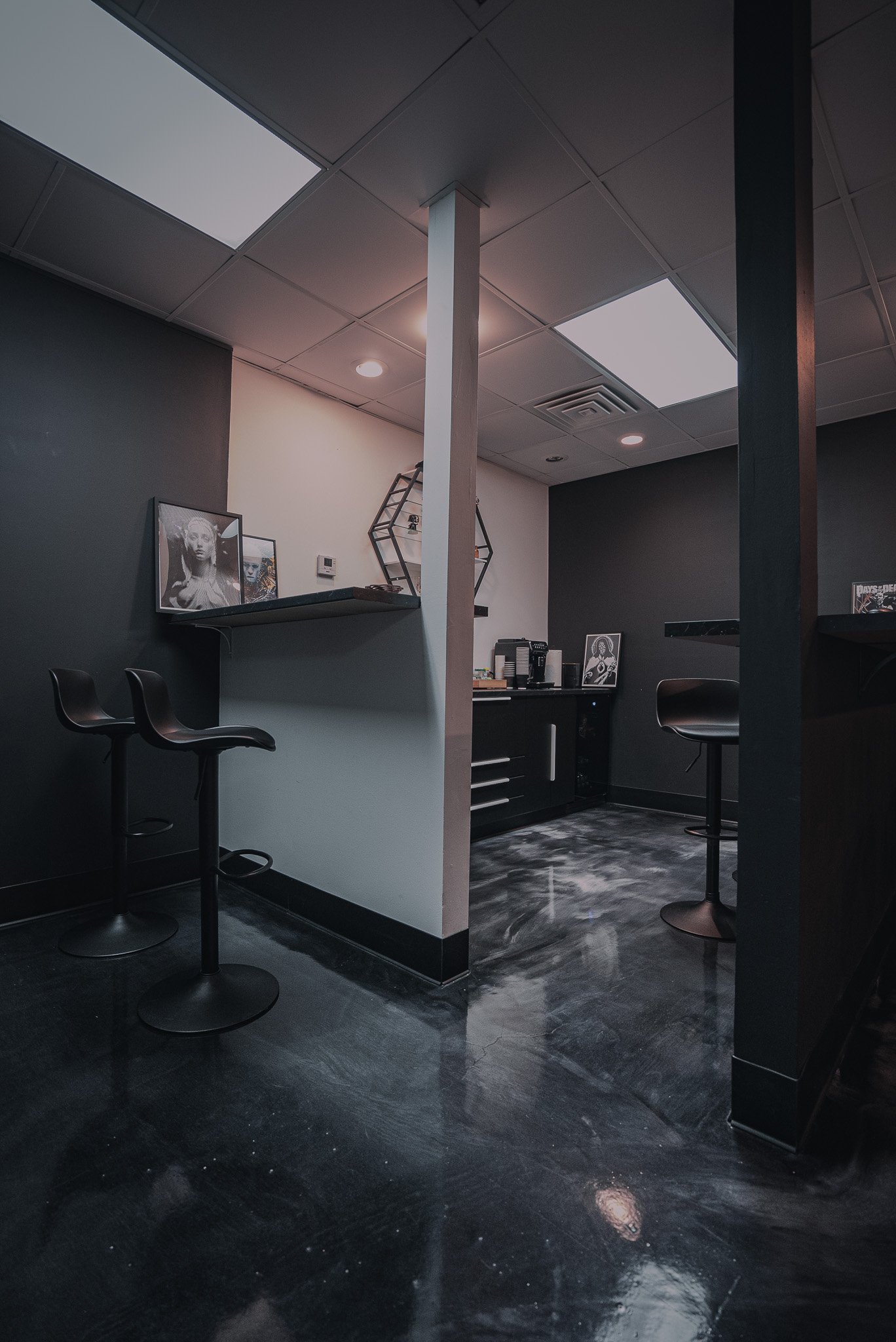 Modern office or lounge space with black and gray color scheme, high stools, art photos on shelves, and a small coffee station on a black countertop.