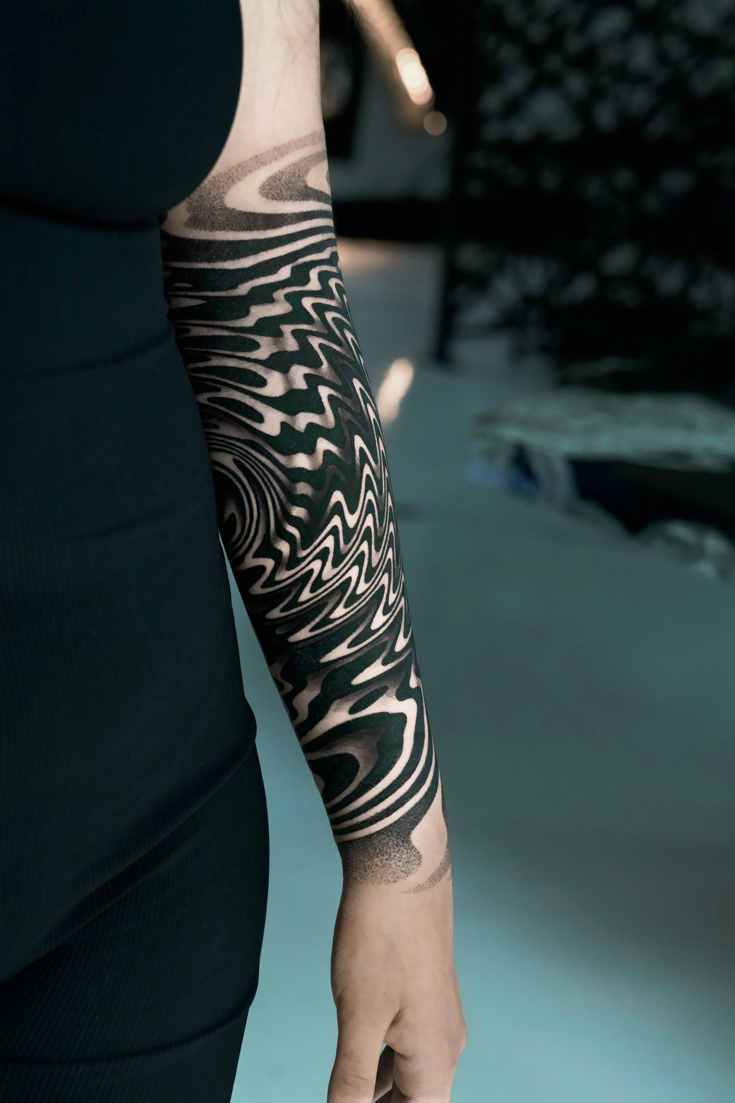 A person with a black and gray patterned tattoo on their arm, wearing a black sleeveless top, standing outdoors with cars and snow in the background.