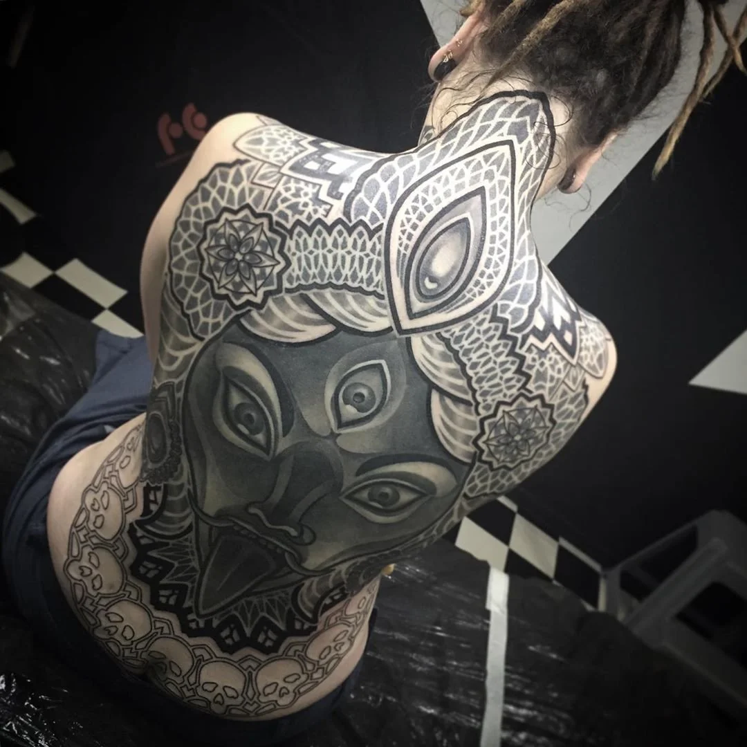 A person with dreadlocks showing their back and shoulder tattoos with intricate black and gray designs, including a large face with multiple eyes, mandala patterns, and other detailed elements.