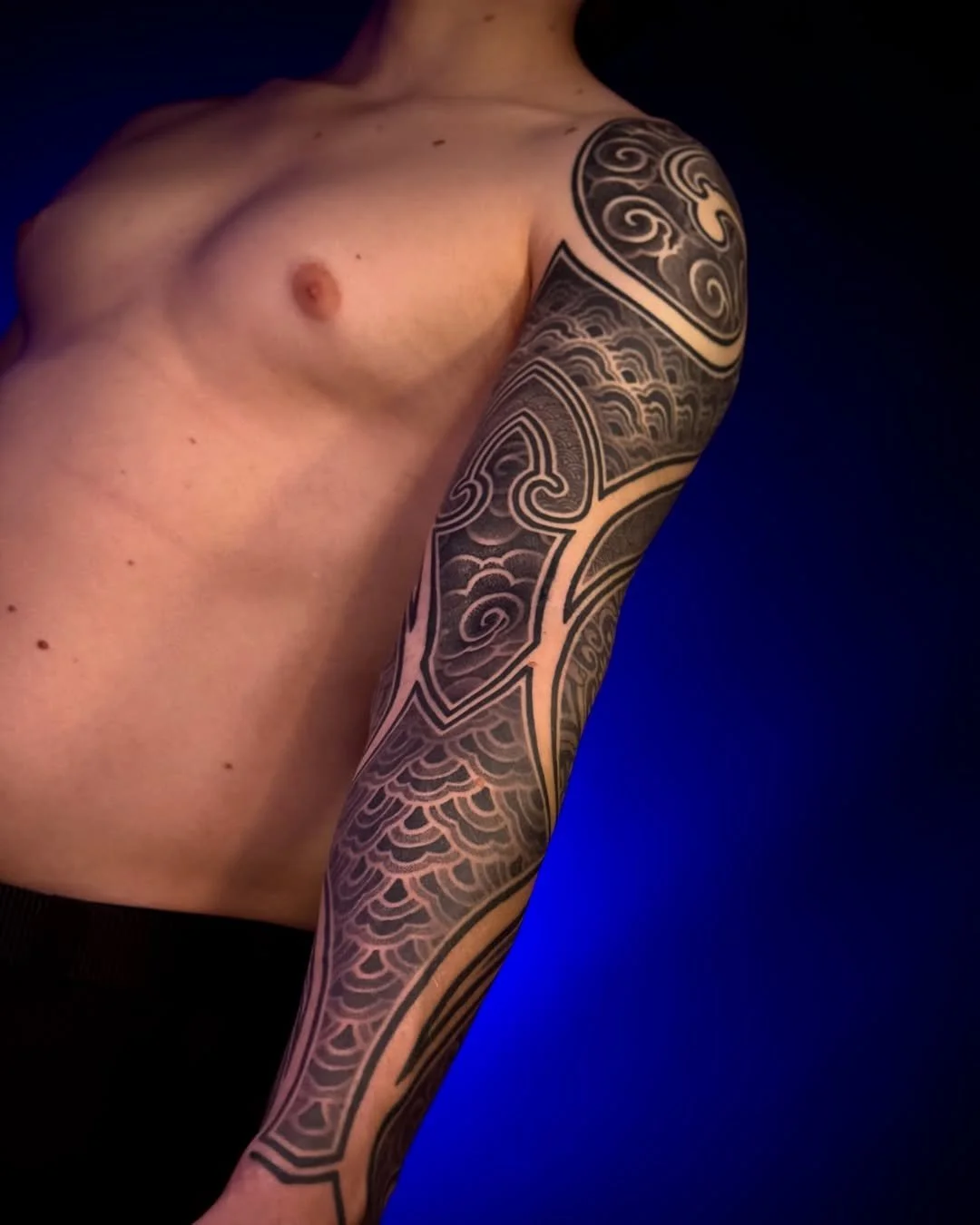 Part of a person's upper body and arm with a black and gray tattoo sleeve against a dark blue background.