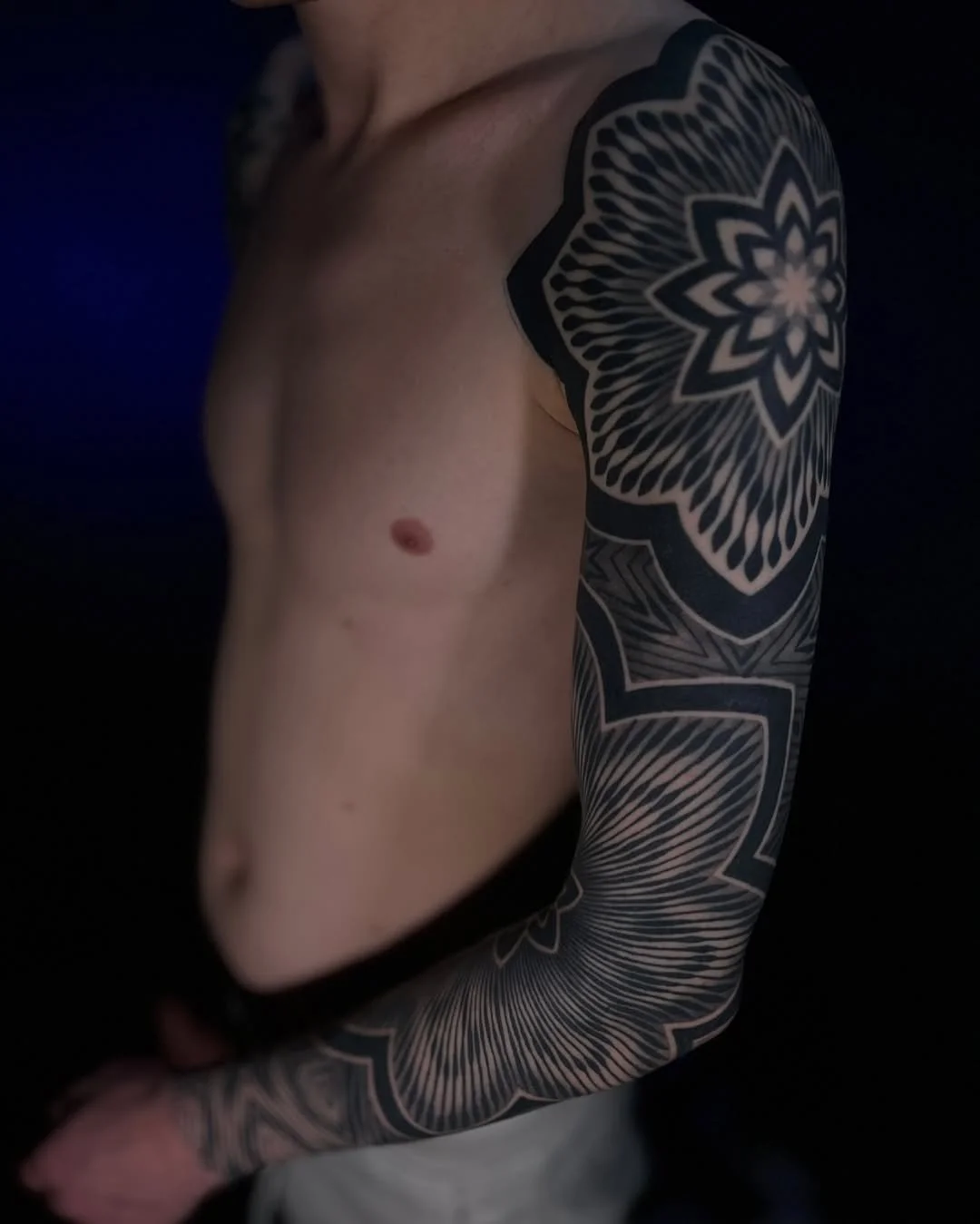 Close-up of a shirtless person with a large black and gray tattoo sleeve on their arm, featuring intricate floral and geometric patterns, against a dark background.