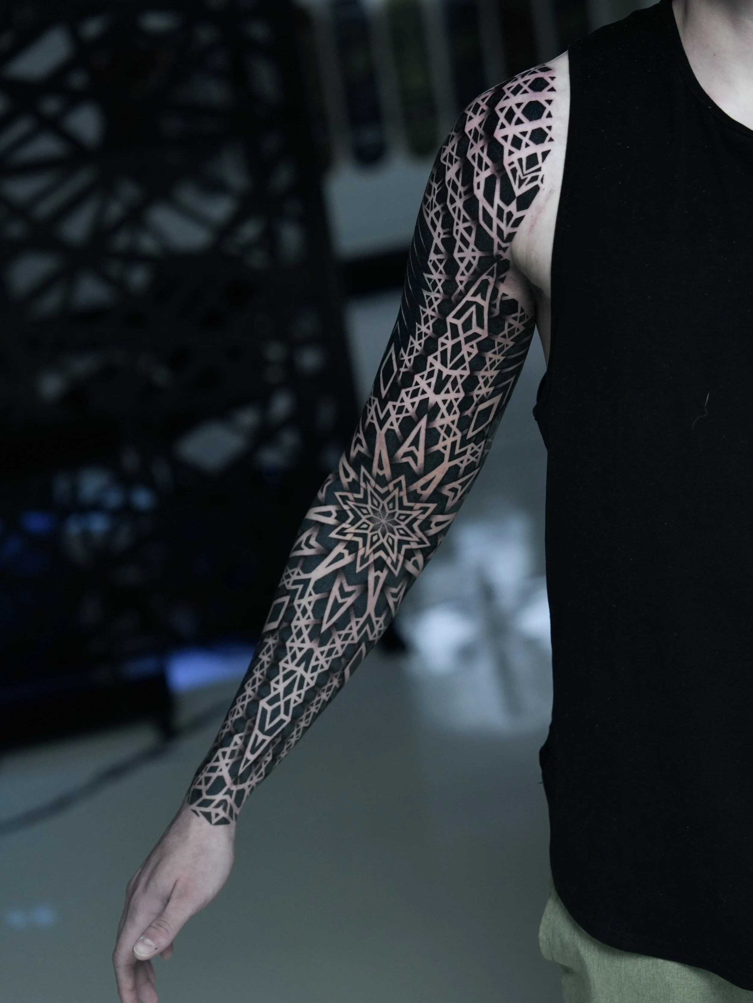 A person with a full sleeve tattoo of intricate geometric patterns extending from shoulder to wrist, wearing a sleeveless black shirt.