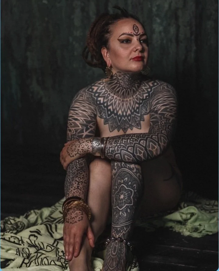 A woman with dark makeup and eye symbols on her forehead, sitting with her legs crossed. She has extensive tattoos covering her arms, legs, and chest, and is wearing jewelry including bracelets and earrings. The background is dark with a hint of gree