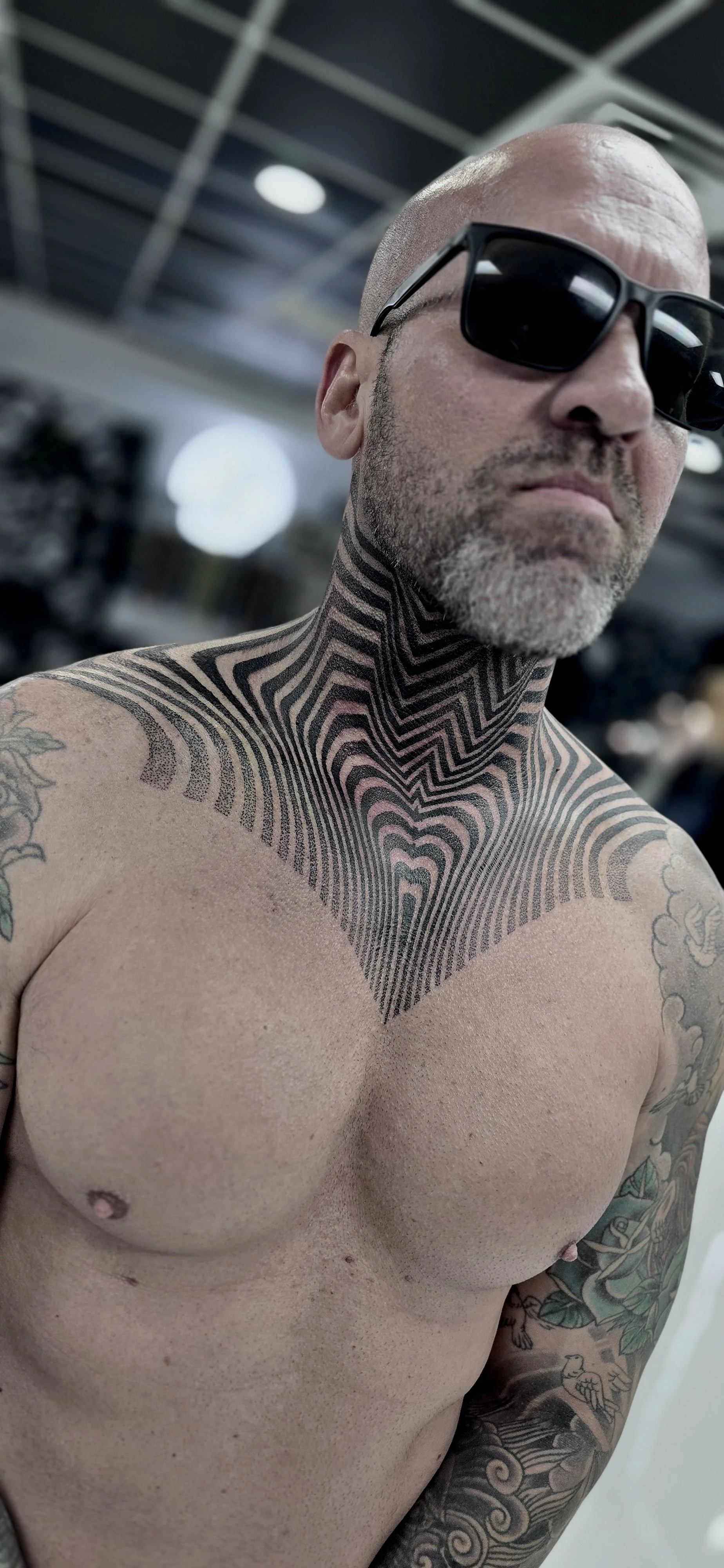 Bald man with tattoos on chest and arm, wearing sunglasses, in an indoor setting with a blurred background.