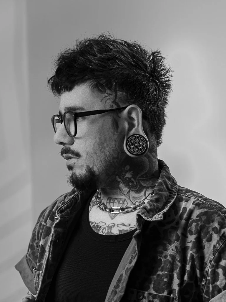 Black and white photo of a man with tattoos, glasses, and large gauge earrings, wearing a camouflage jacket and chain necklace, with a side profile view.
