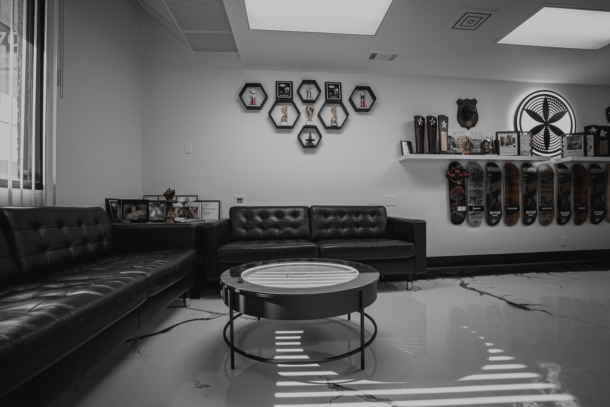 A black and white photo of a waiting room with leather couches, a round coffee table, skateboard awards on hanging plaques, a shelf with trophies, framed photos, sports memorabilia, and a patterned decorative wall piece.