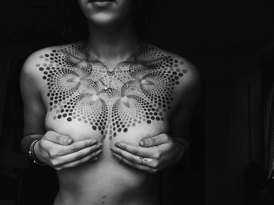 A woman covering her breasts with her hands, showcasing an intricate geometric tattoo on her chest and shoulders in black and white.