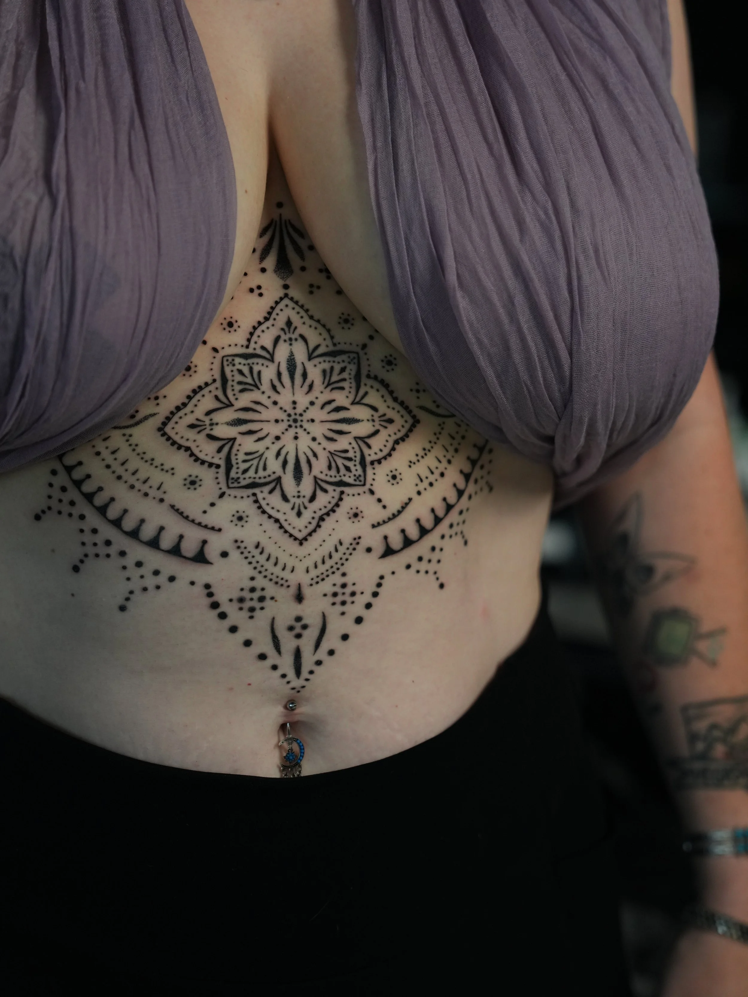 Close-up of a woman's torso with a large, intricate black tattoo on her chest, a belly button piercing, and tattoos on her arms, wearing a purple top with a V-neck.