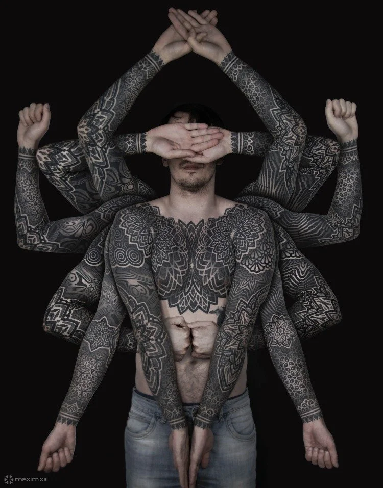Multiple arms and hands, some tattooed, are positioned around a shirtless man with extensive tattoos, against a black background.