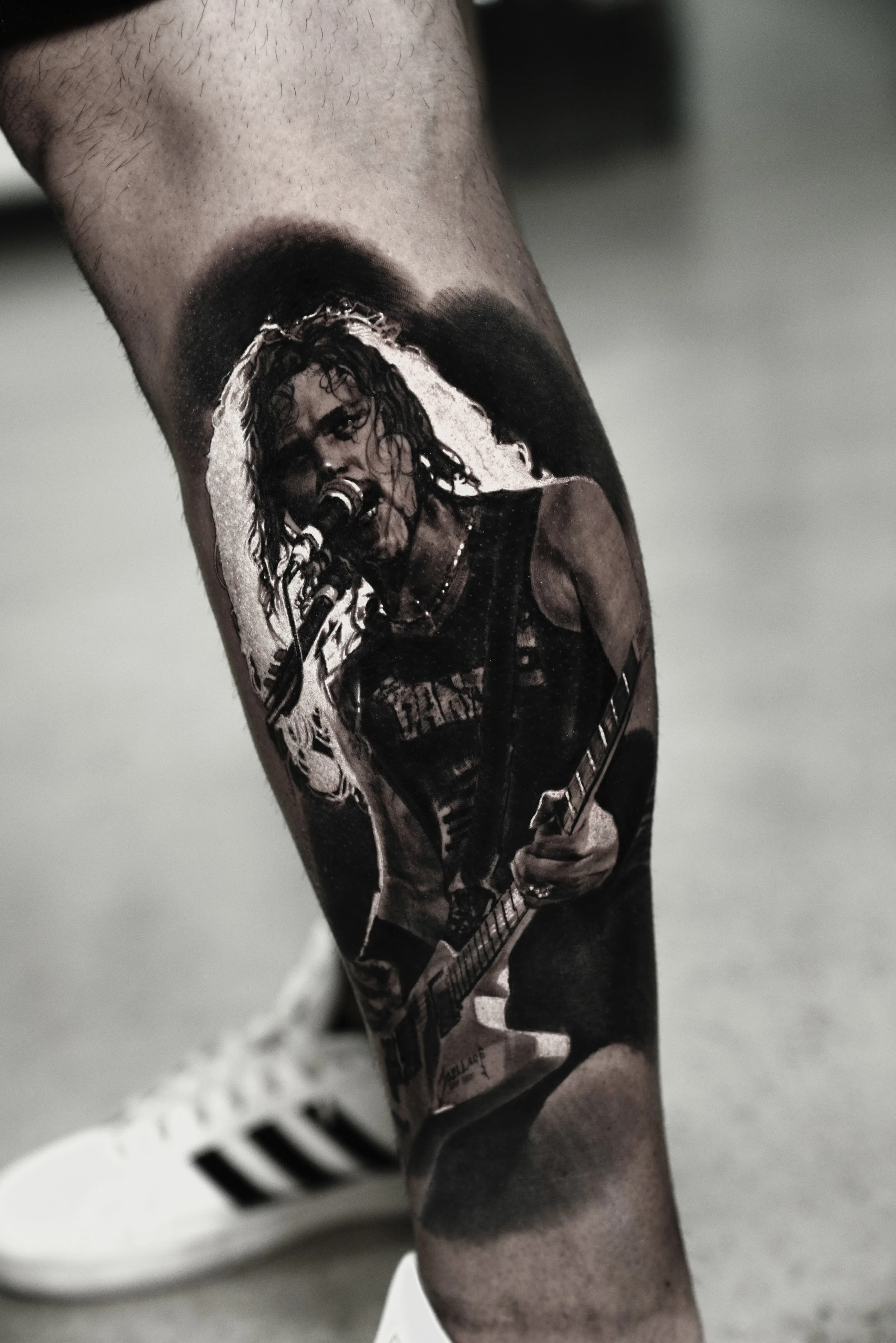 A close-up of a person's leg with a black and white tattoo of a musician singing into a microphone, wearing a sleeveless shirt, holding an electric guitar, with a dark background.