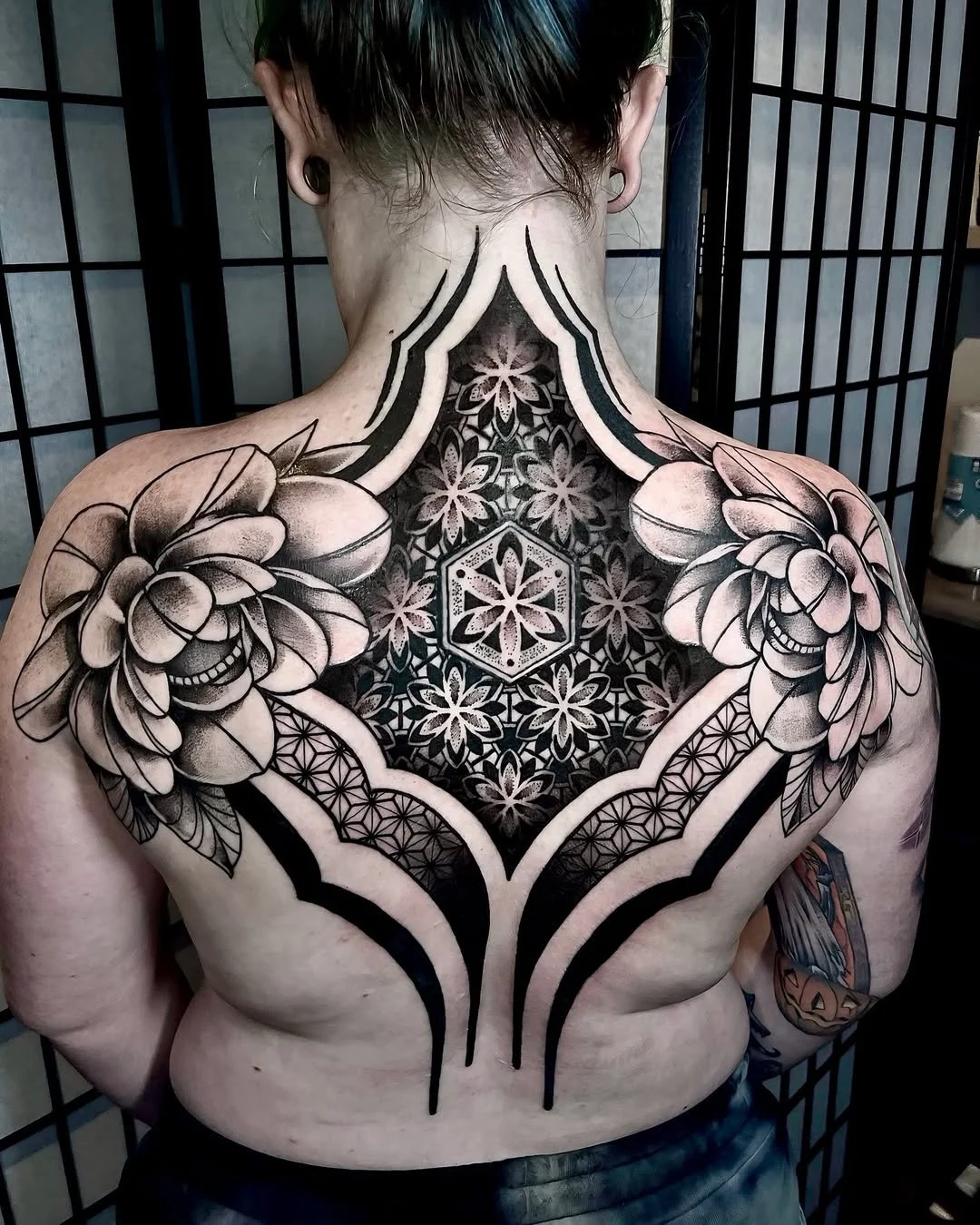 Black and gray geometric and floral tattoo covering the person's chest and shoulders, featuring large flowers, honeycomb patterns, and bold lines.