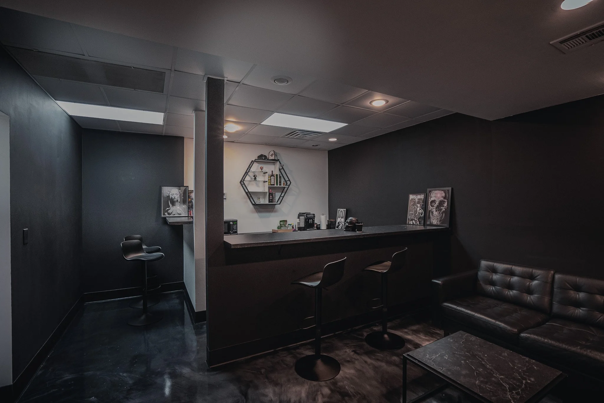 A modern, dimly lit lounge or bar area with black walls, a black leather sofa, a dark marble coffee table, and a bar counter with two black barstools. Shelves with artwork and bottles are on the wall behind the bar.