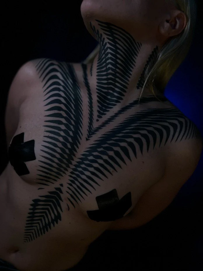 A person with intricate black body paint or tattoos resembling fern leaves across their chest and neck, with black tape covering their nipples, in a dark setting.