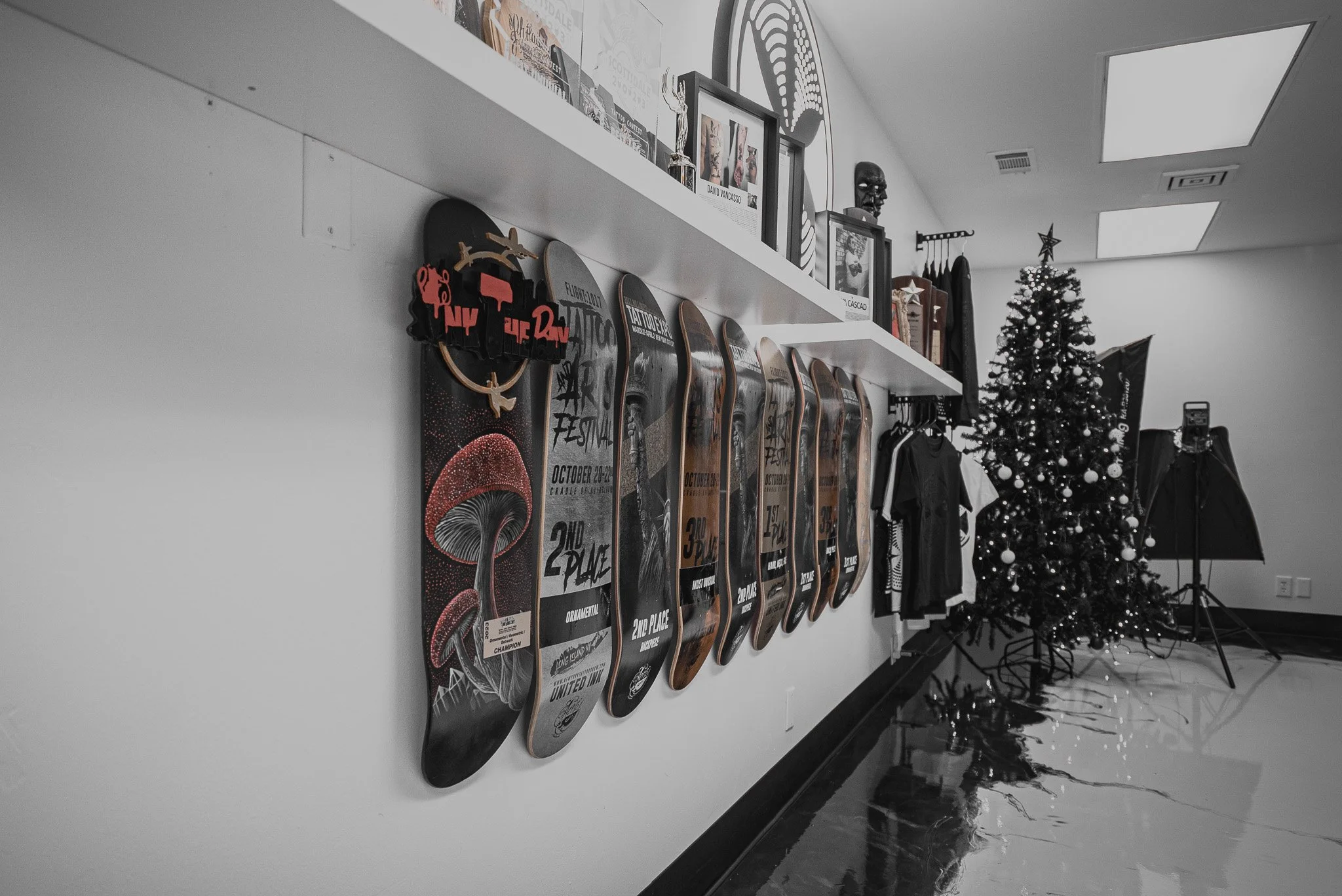 A white wall with skateboards hanging in a row, framed photos and artwork on a shelf above, a Christmas tree with lights and ornaments, and photography studio equipment including a light and umbrella in the background.