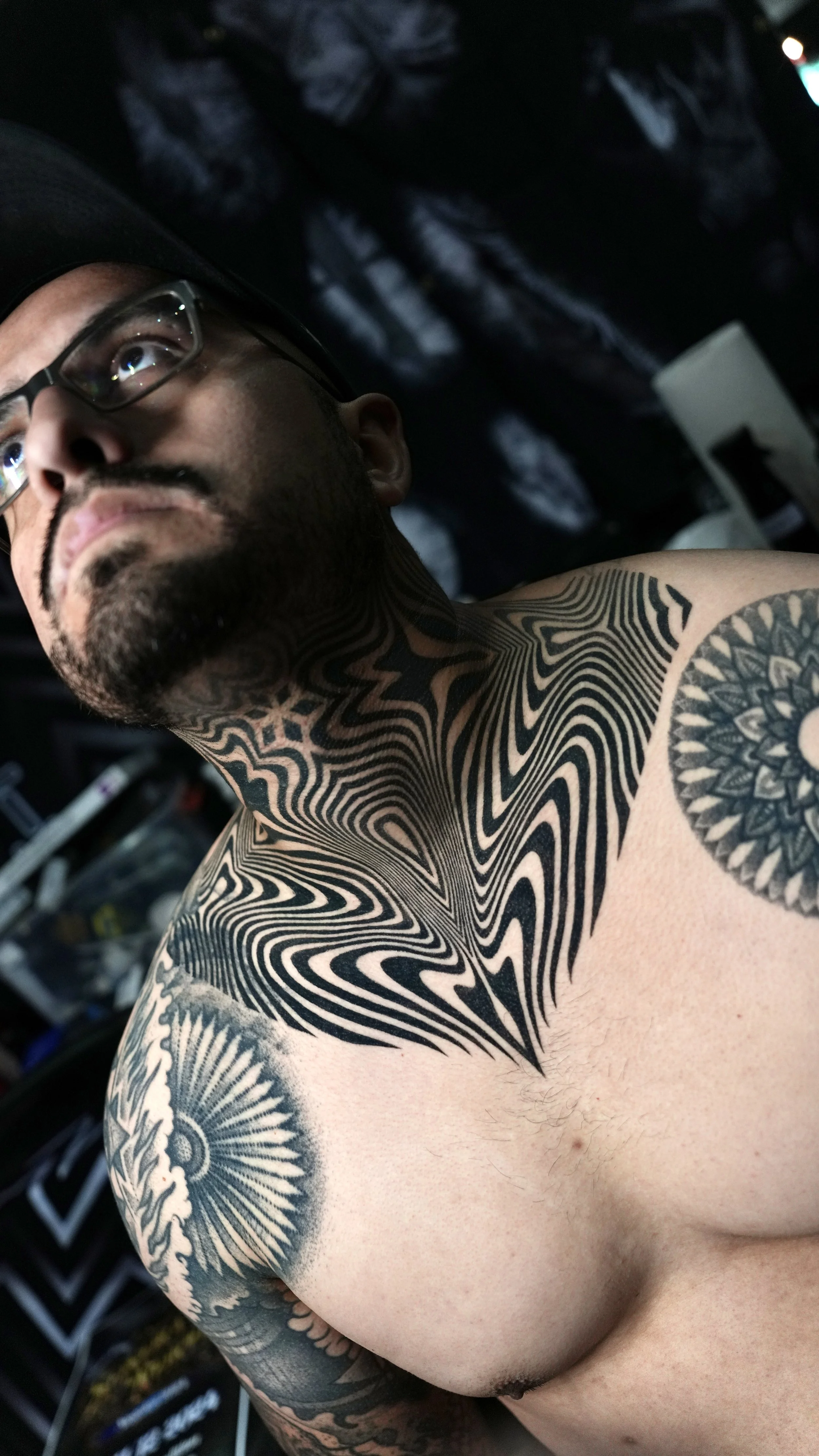 A shirtless man with glasses and facial hair, looking to the side, featuring intricate black and gray tattoos on his neck, shoulder, and chest, including geometric patterns, spiral designs, and a sun motif.