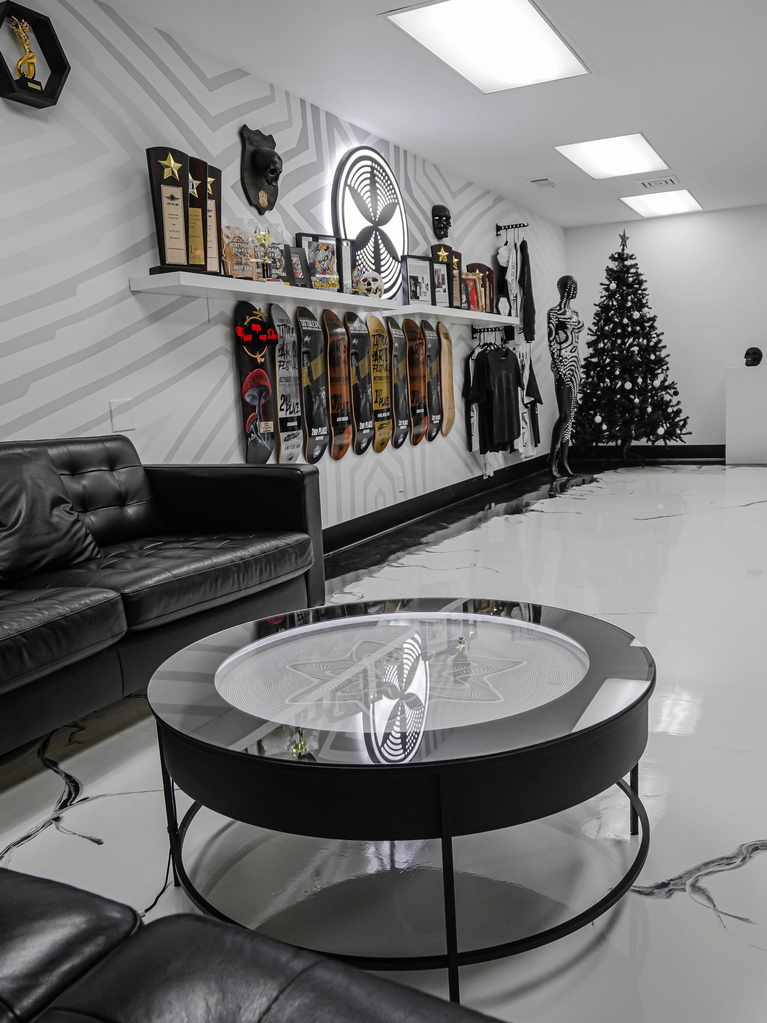 Interior of a modern lounge with black leather sofas, a round black coffee table with a reflective top, skateboard decks on the wall, a decorated Christmas tree, and various art pieces and memorabilia on a shelf.