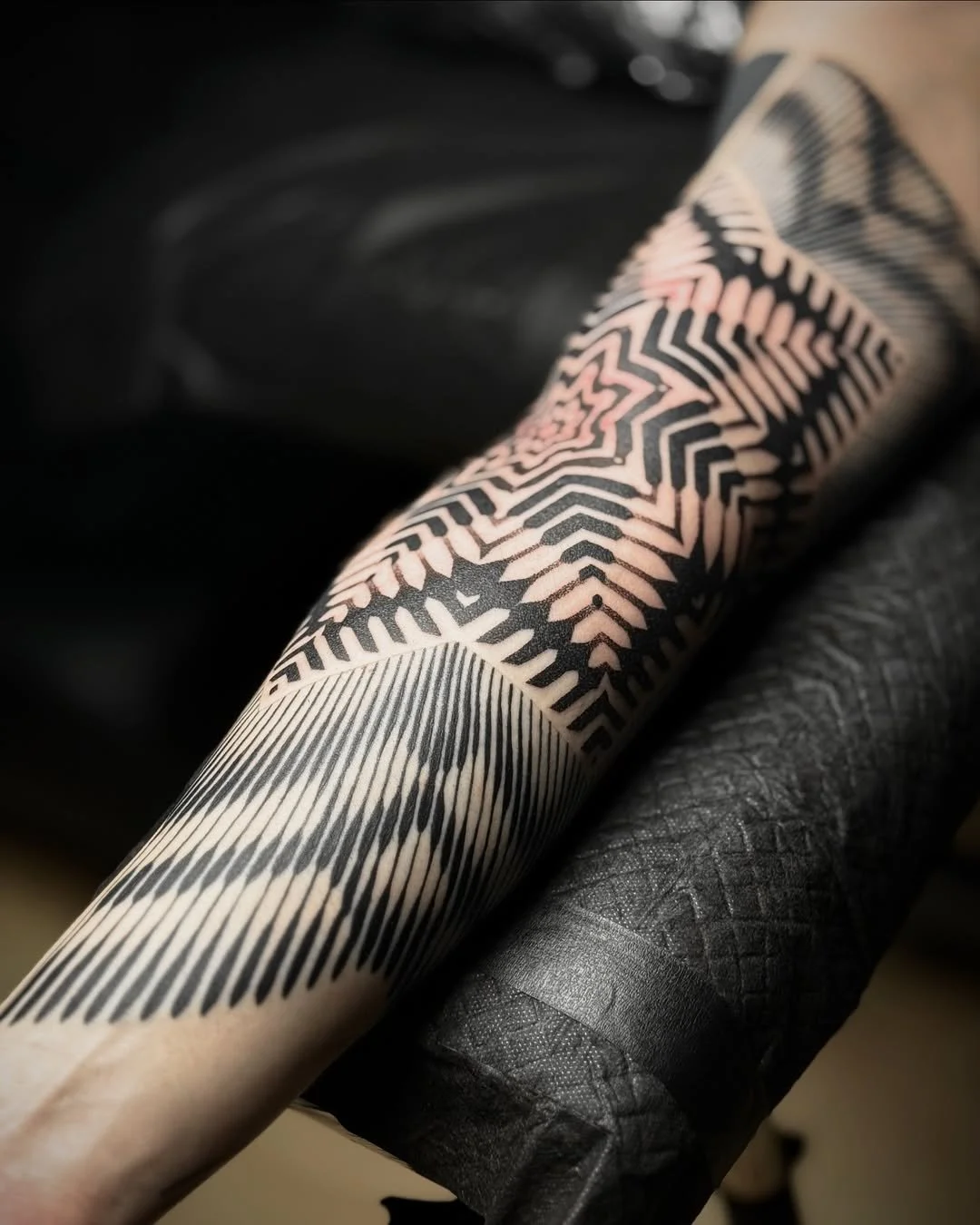 A close-up of a forearm with a black geometric tattoo featuring radial lines and zigzag patterns.